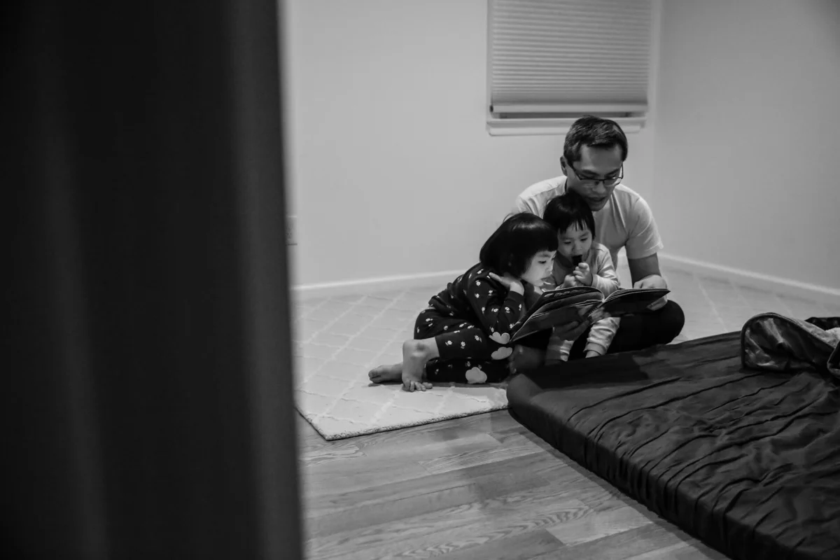 Man with glasses sits on rug with two children in pajamas, reading a book