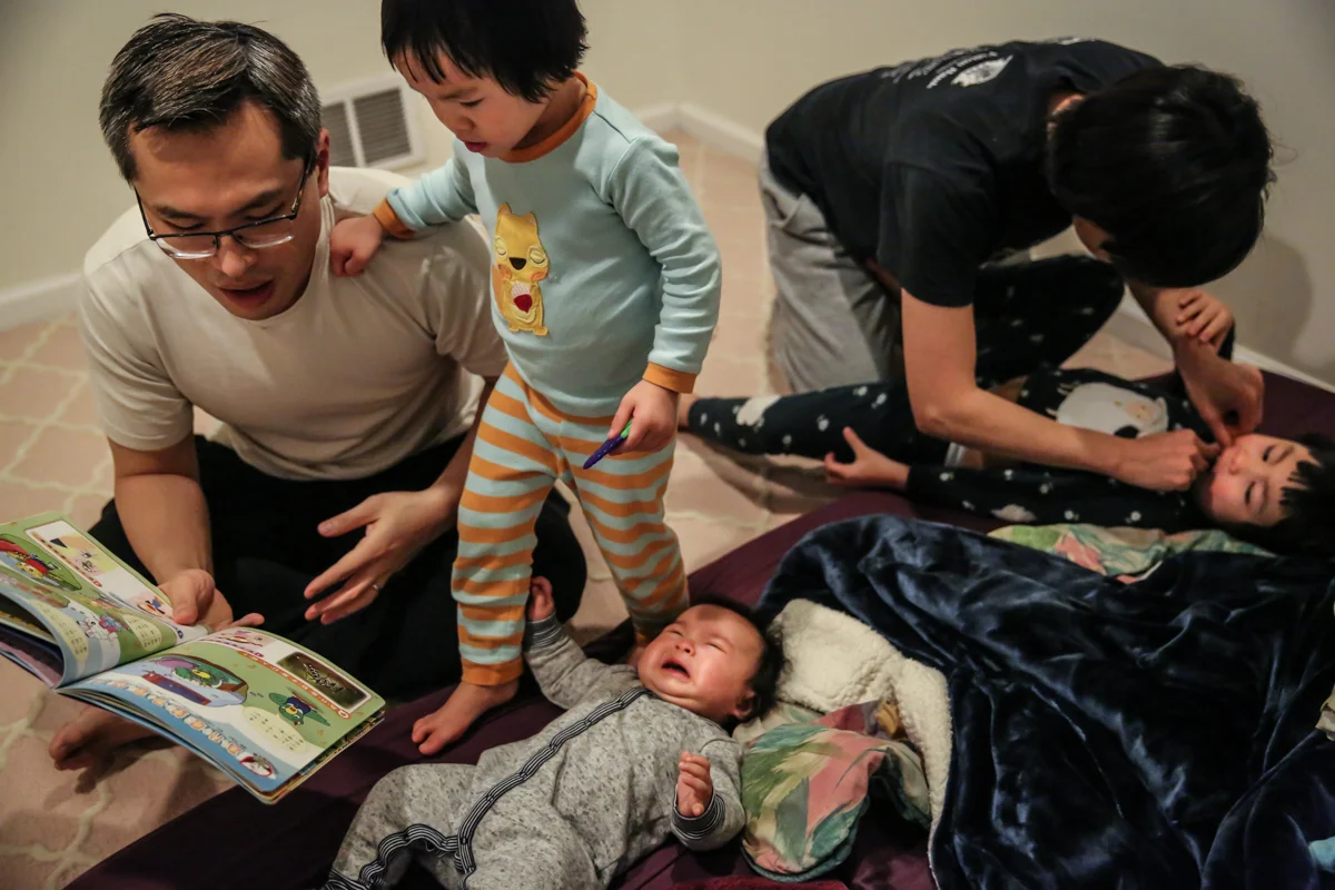 Baby cries on bed while man with glasses reads book and girl in pajamas walks over baby. Woman flosses other girl’s teeth on bed