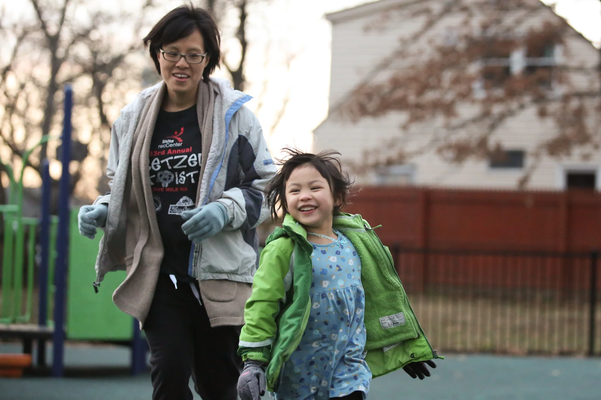 Girl in blue dress and green jacket smiling and running while woman wearing glasses runs behind girl
