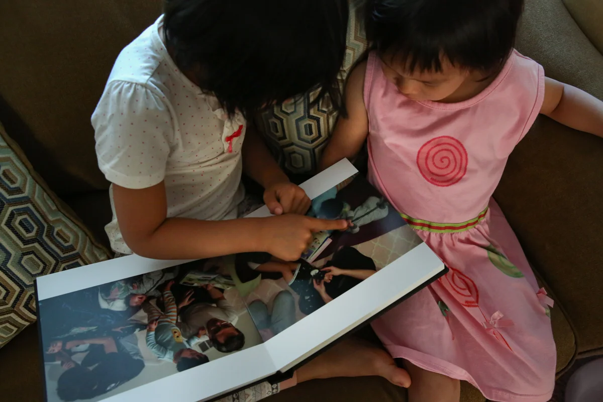 2 girls sitting on a couch looking and pointing at photo album. One girl is in a white and red polka dot shirt, the other girl is in pink dress with red swirl.
