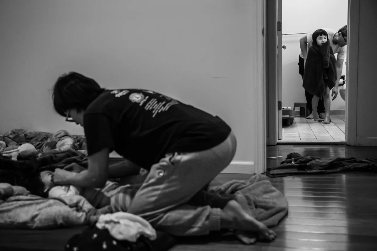 Woman flosses girls teeth on floor while girl stands in bathroom doorway wrapped in towel