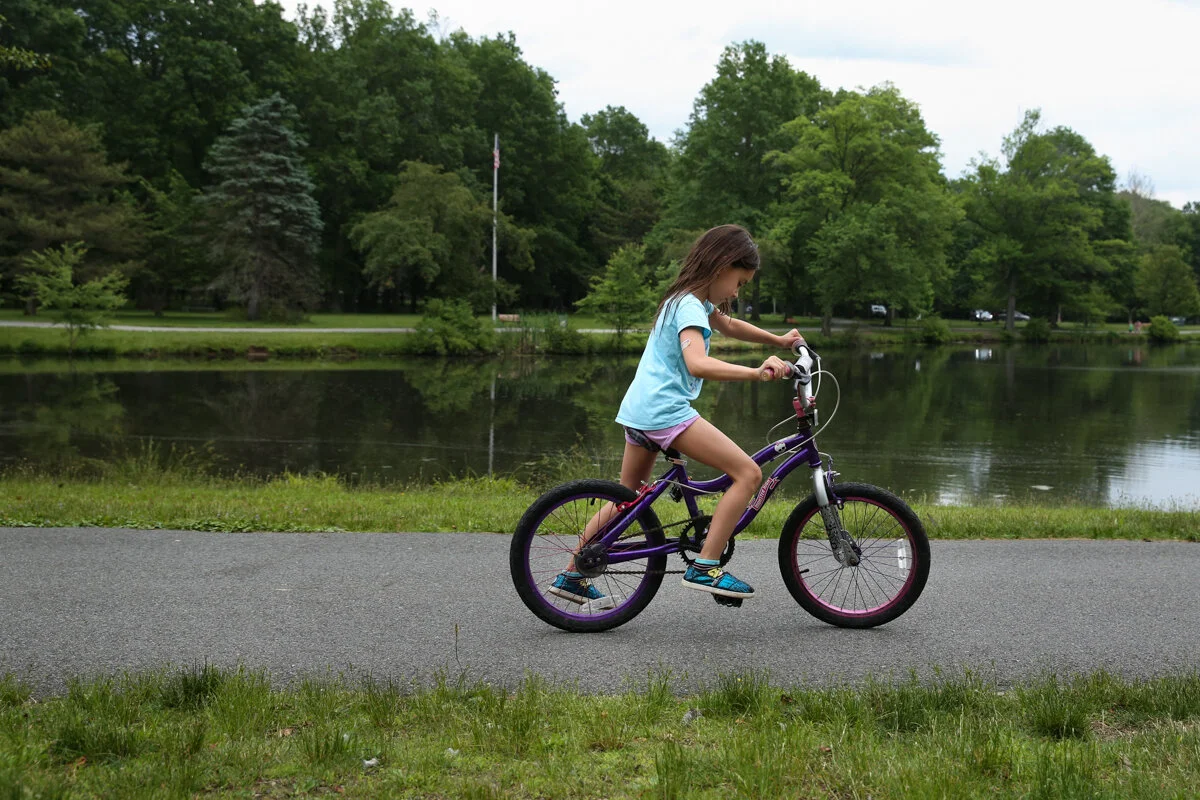 Girl rides bike on asphalt path with pond and trees in background
