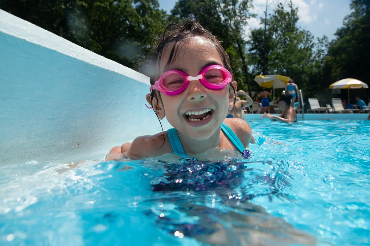 Girl with pink goggles and missing lower tooth smiles while in a swimming pool