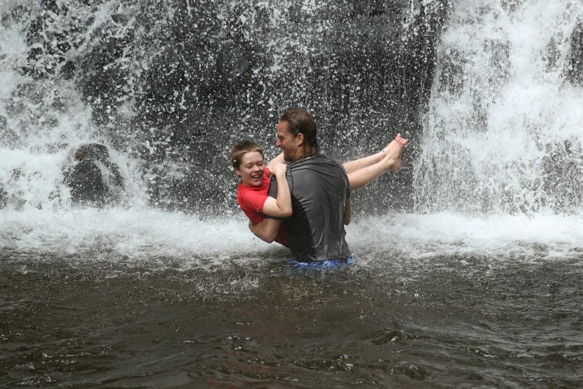 Man carries boy wearing red shirt into a waterfall