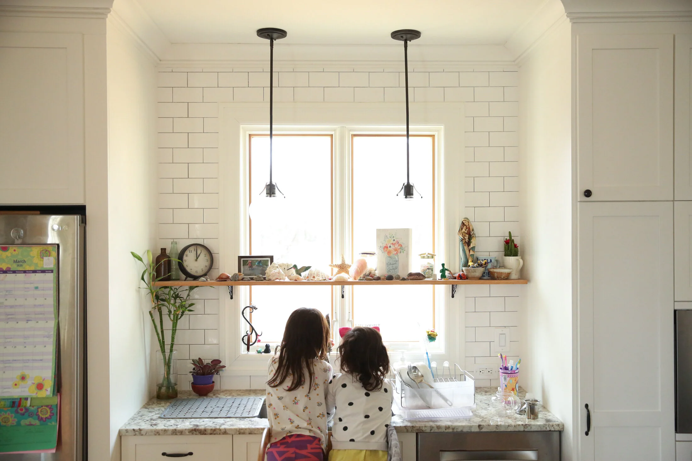 Two dark haired children stand in front of a sink window with their backs facing the camera.