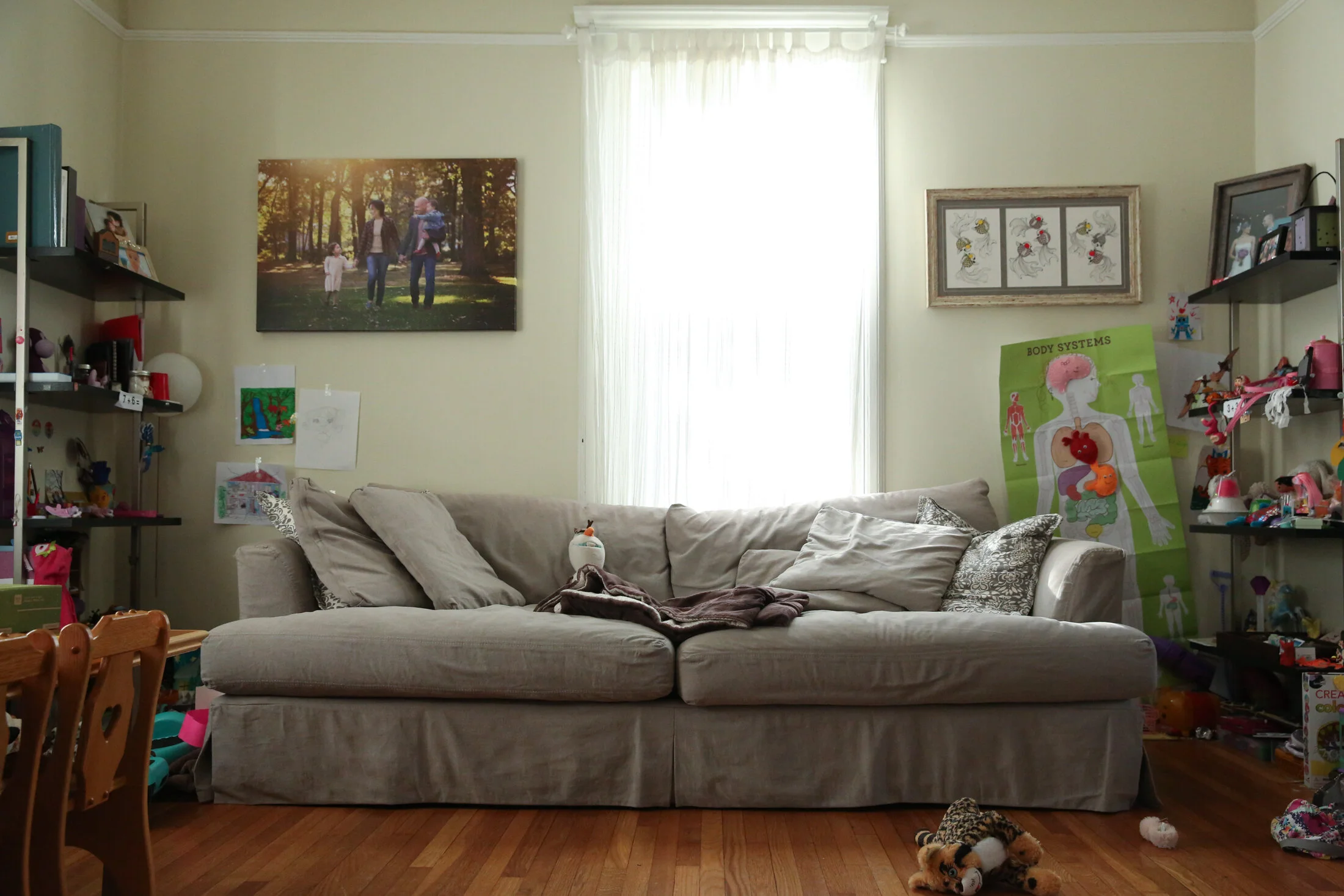 A grey couch is situated in front of a window. Toys shelf flank the left and right of the couch while photos hang on either side of the window.