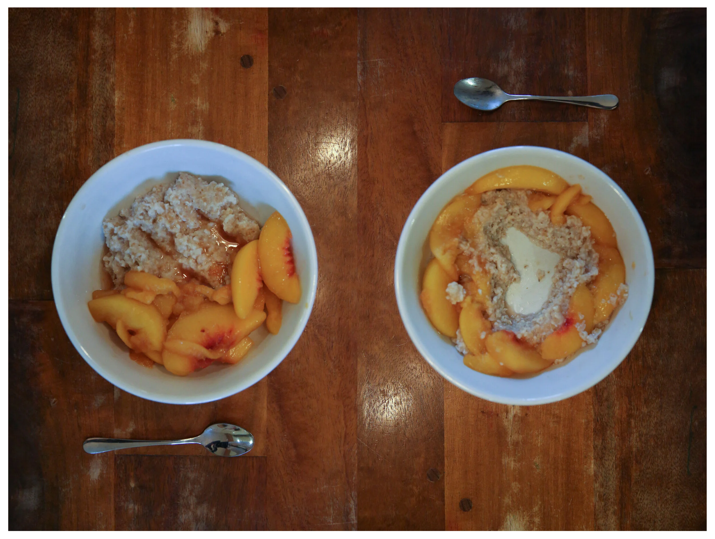 Two bowls of oatmeal and and cooked peaches on a wooden table. The one on the left has a heart shape eaten out of the oatmeal.