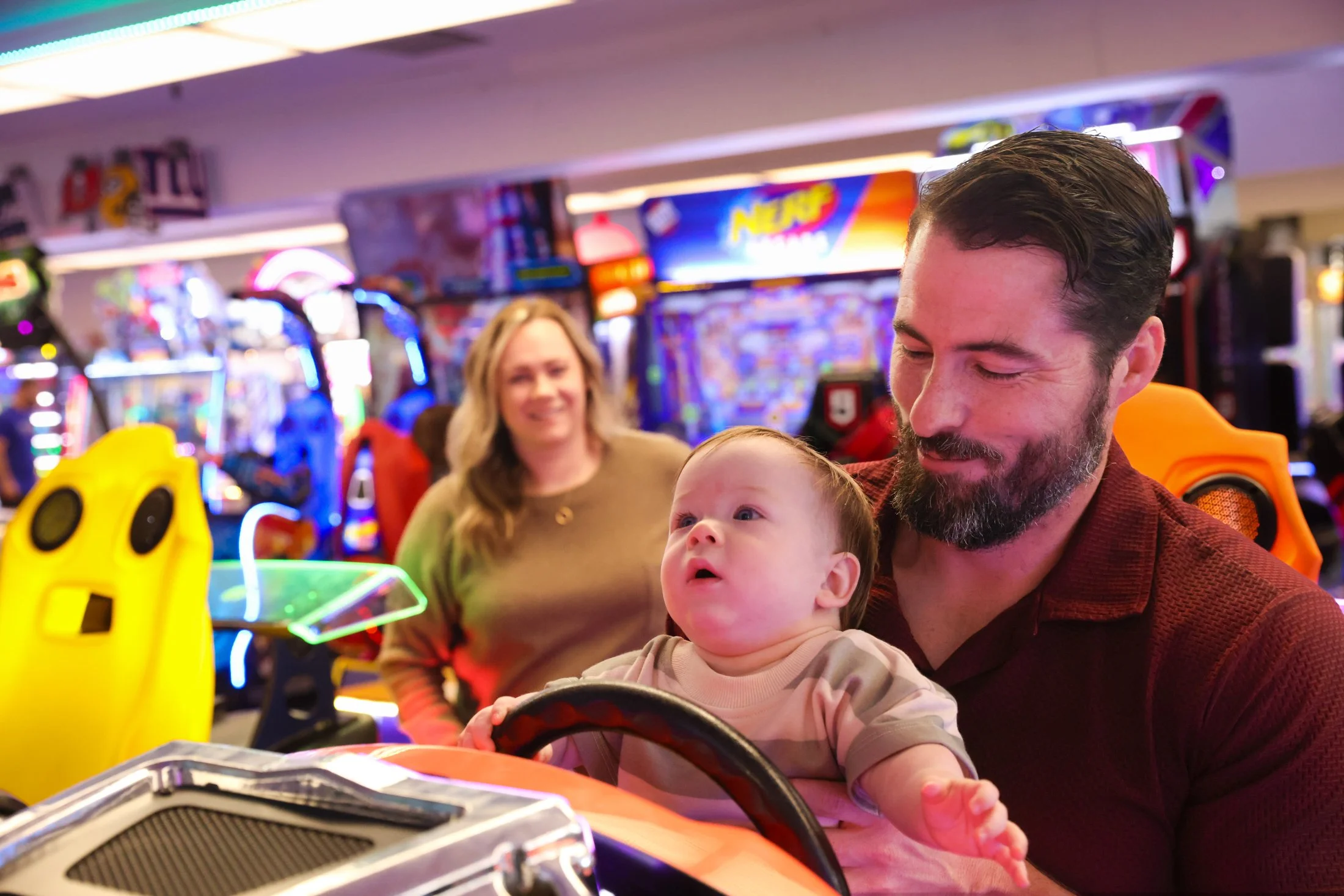 a father holds his baby in front of a steering wheel in a video arcade, mother looks from behind