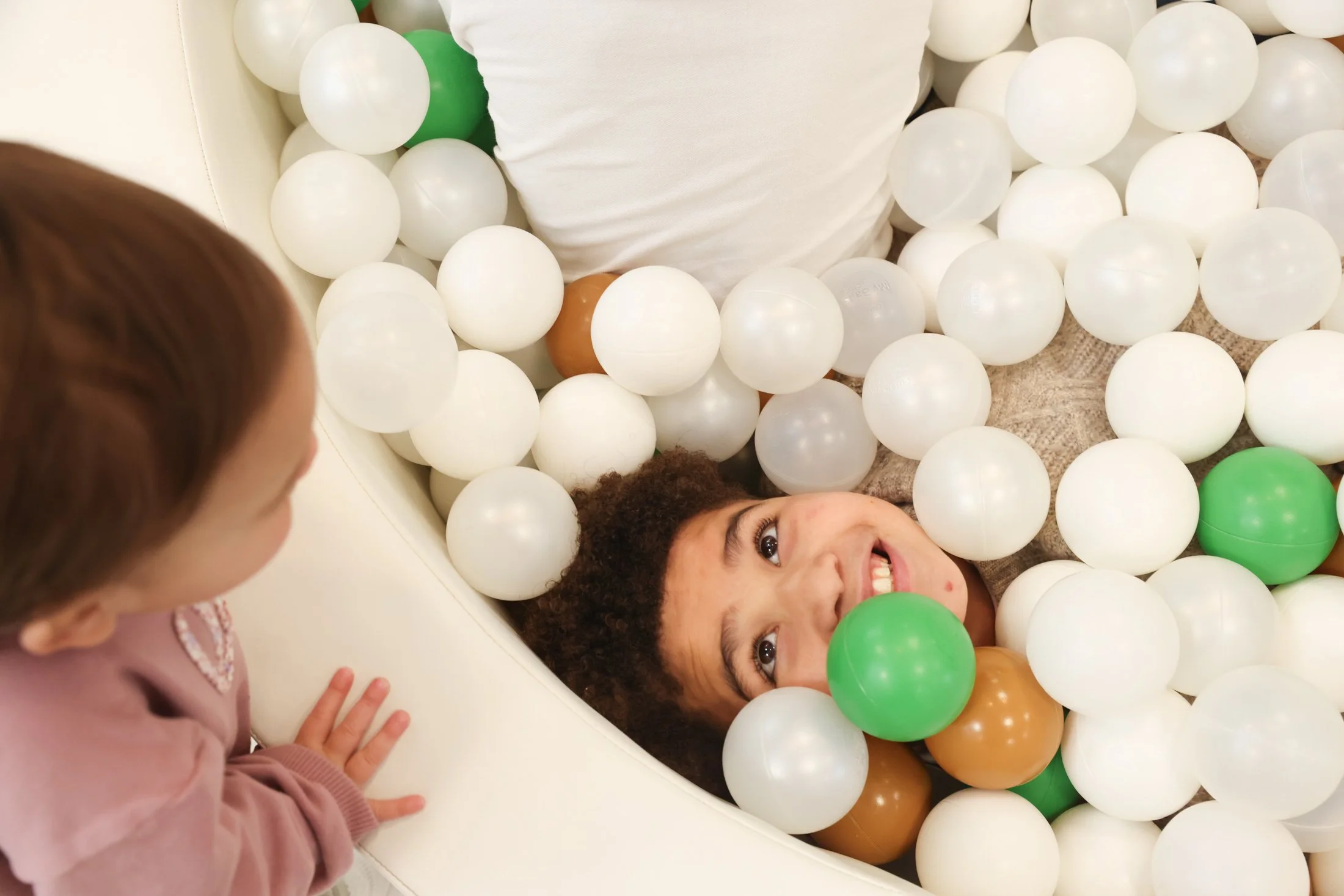 face of boy looking up from a ball pit, looking at a young toddler peering into the ball pit