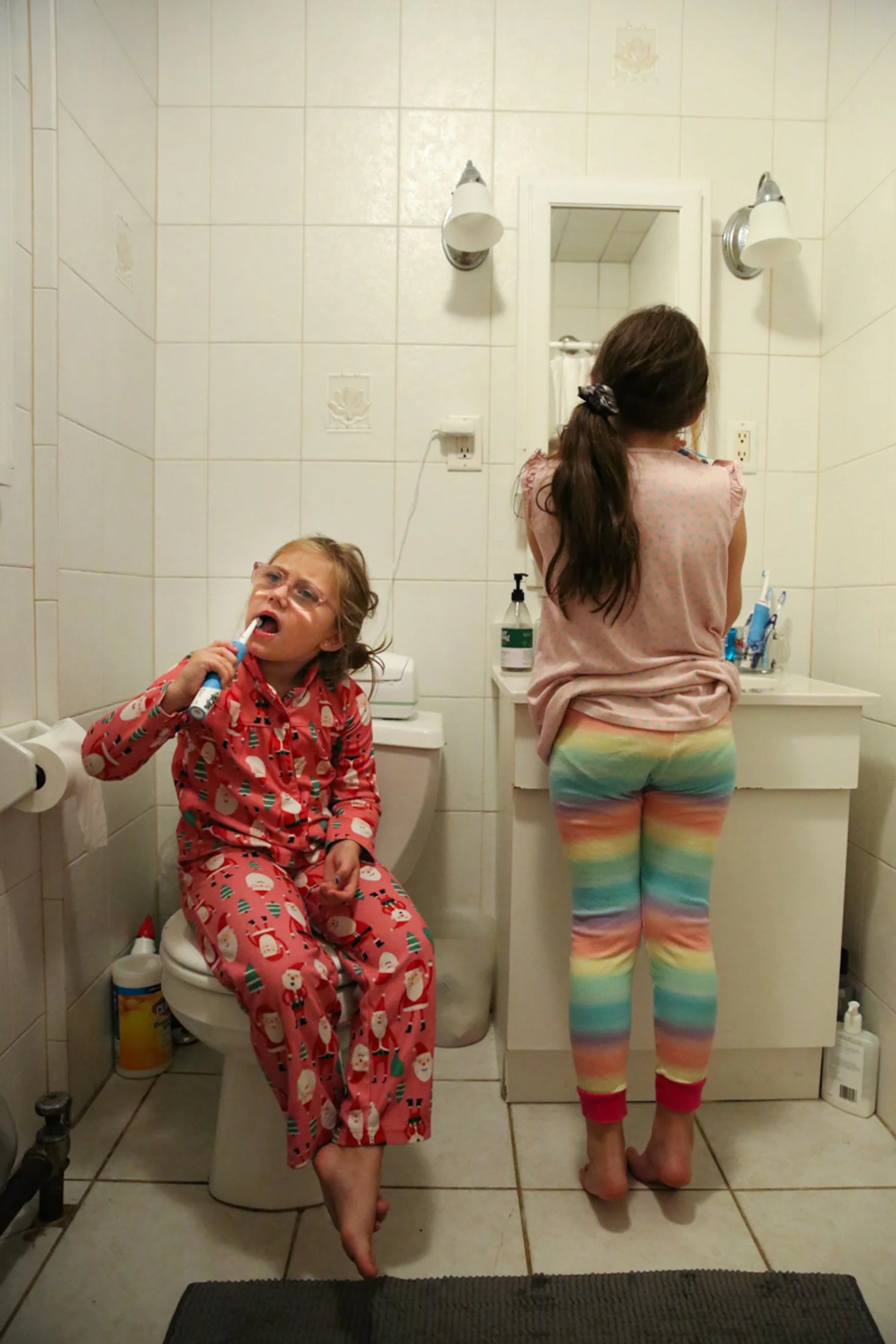 2 girls in pajamas brushing their teeth. one girl stands back towards the camera, 2nd girl is sitting on toilet seat while brushing teeth