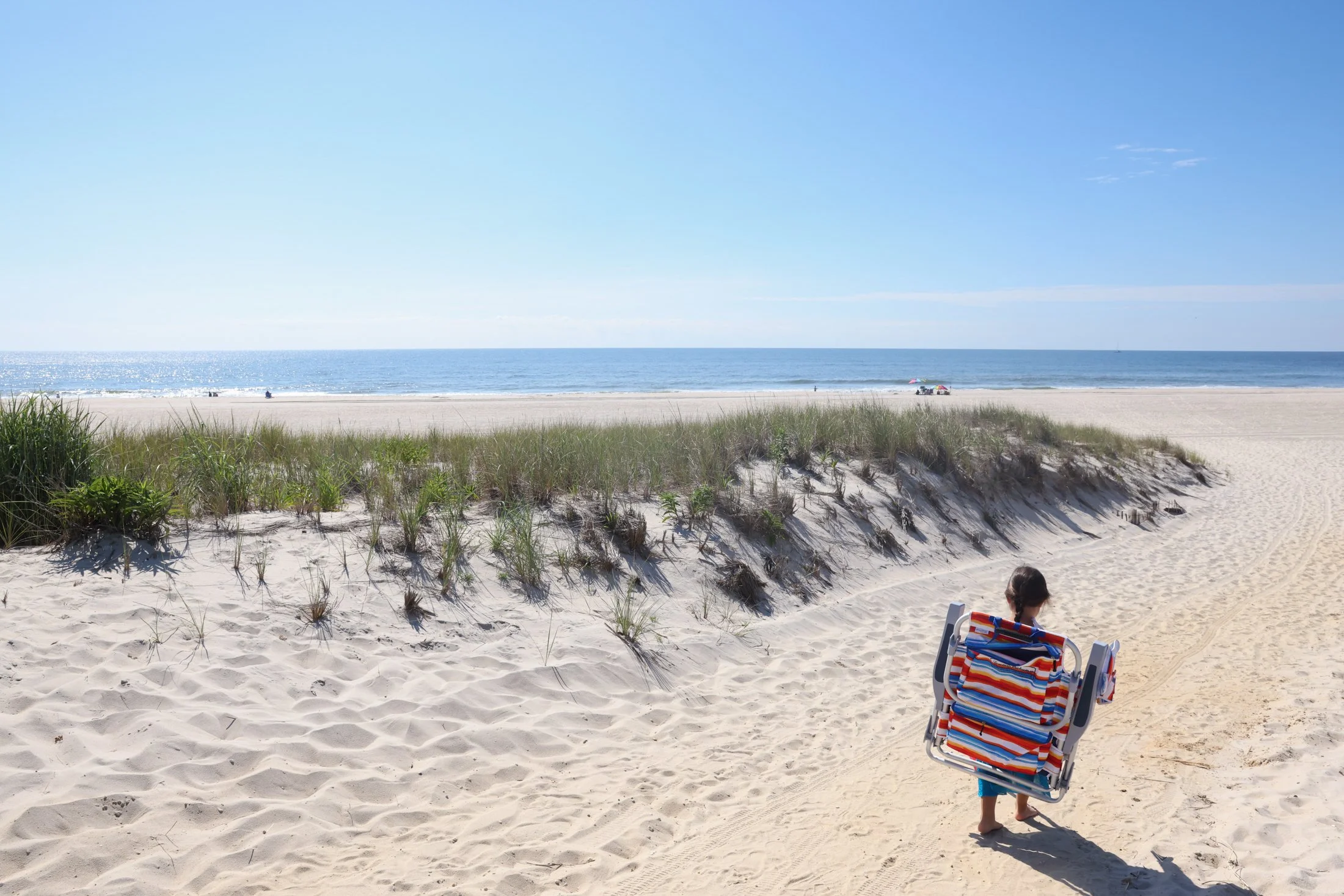 young child with folding chair on their back walking towards the beach