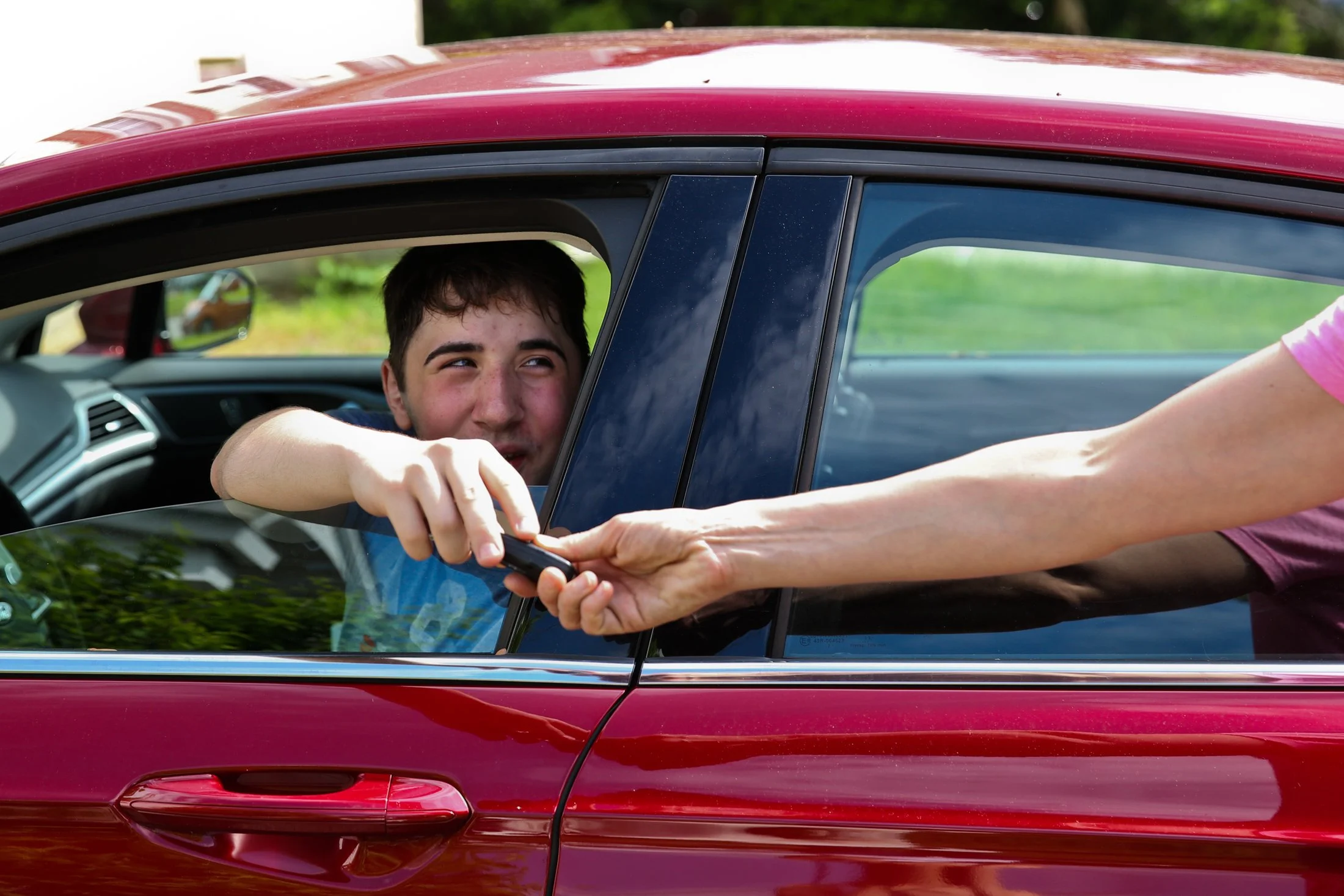 an adult arm passes keys to a teenage boy in driver seat of a red car, through open window