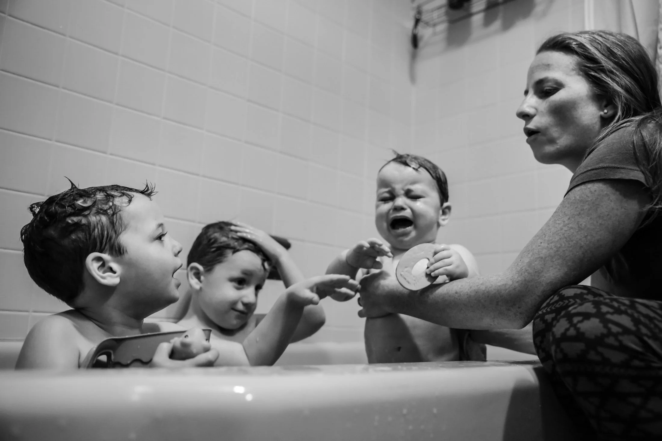 a mother bathes her 3 young children in a bathtub. the youngest child is crying, one child is talking, one child is scratching their head