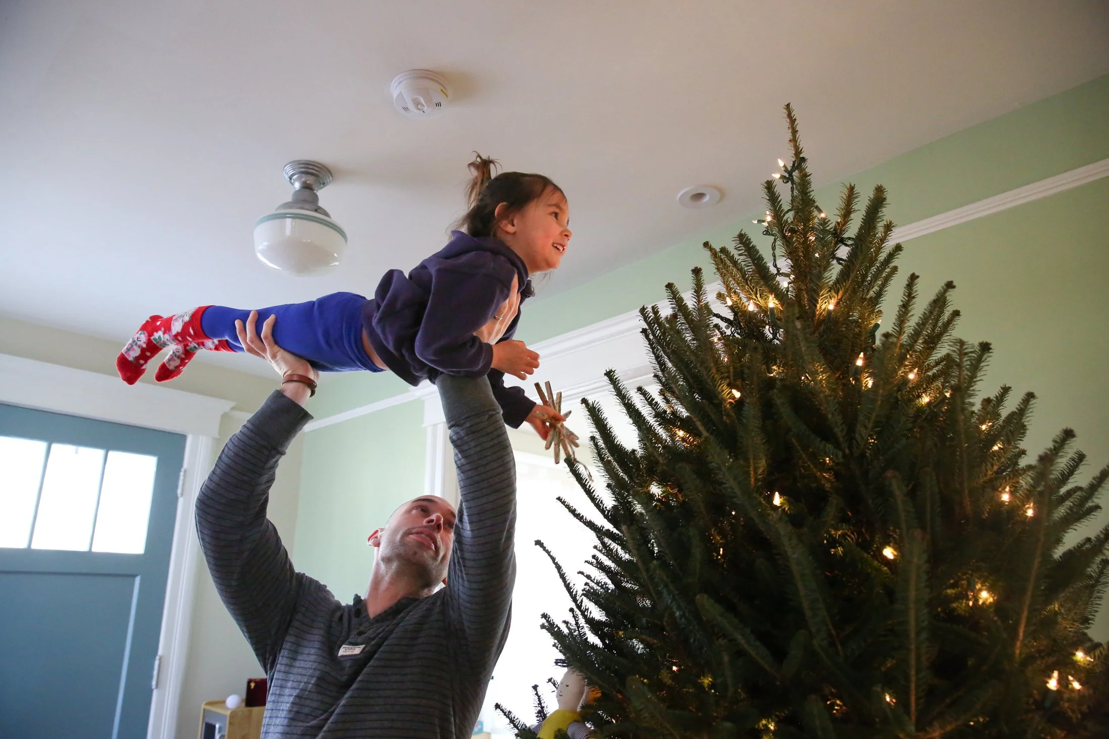 father holds daughter up in the air like Superman as she attempts to place a star on top of a lit Christmas tree