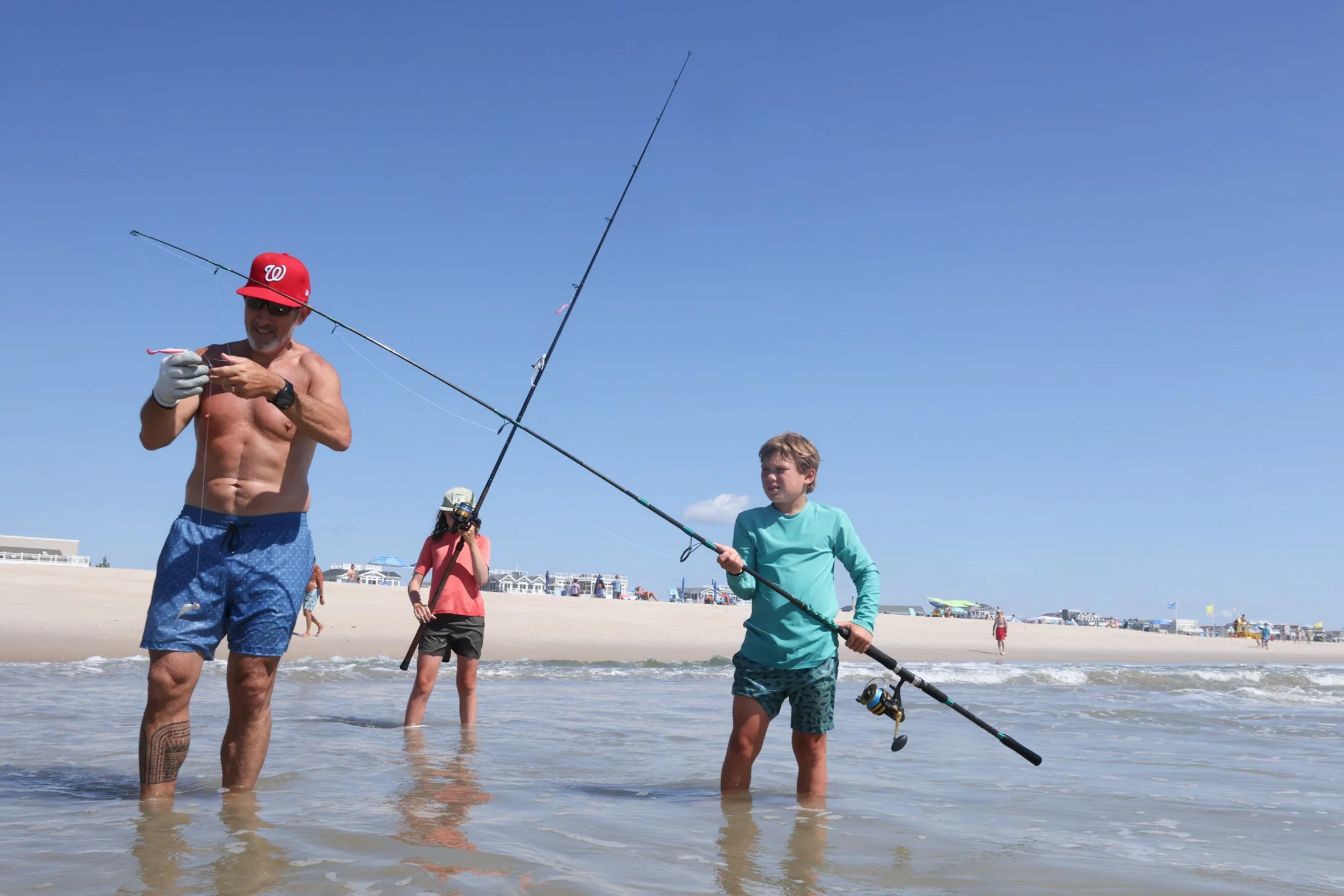 a man and 2 school aged children standing at low tide on the beach. shirtless man is wearing blue swim trunks and red cap while trying to untangle a fishing line that 1 child is holding. the second child stands behind holding their own fishing pole