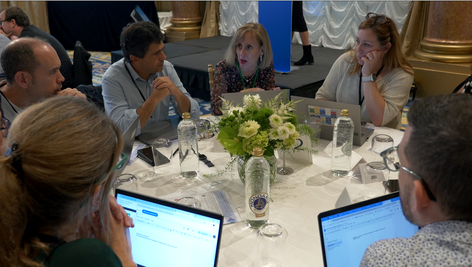 A group of people engaged in a discussion around a round table at a conference. There are laptops, water bottles, glasses, and a floral centerpiece on the table.