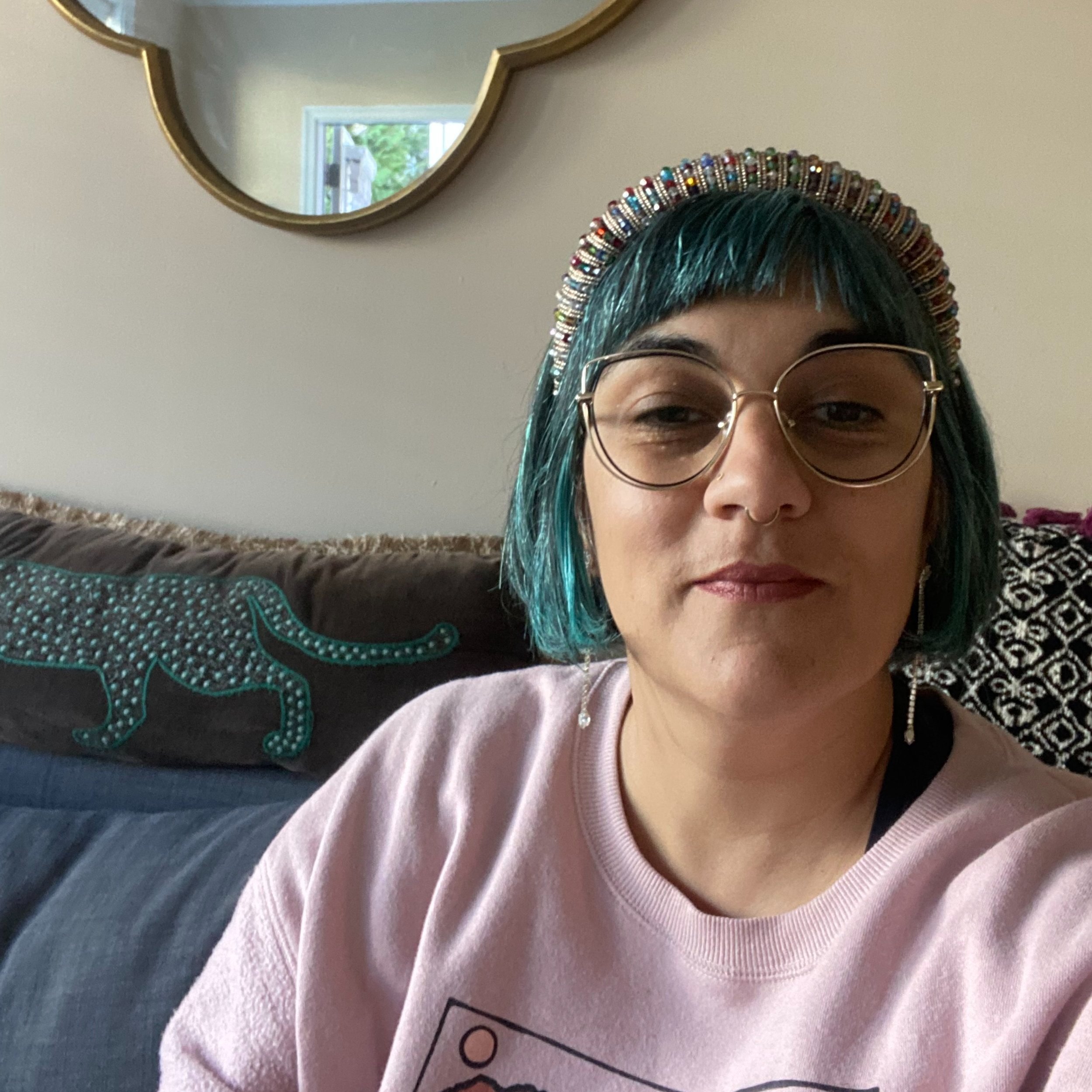Person with teal-colored hair and glasses, wearing a beaded headband, in an indoor setting with a wall mirror and an open window in the background.