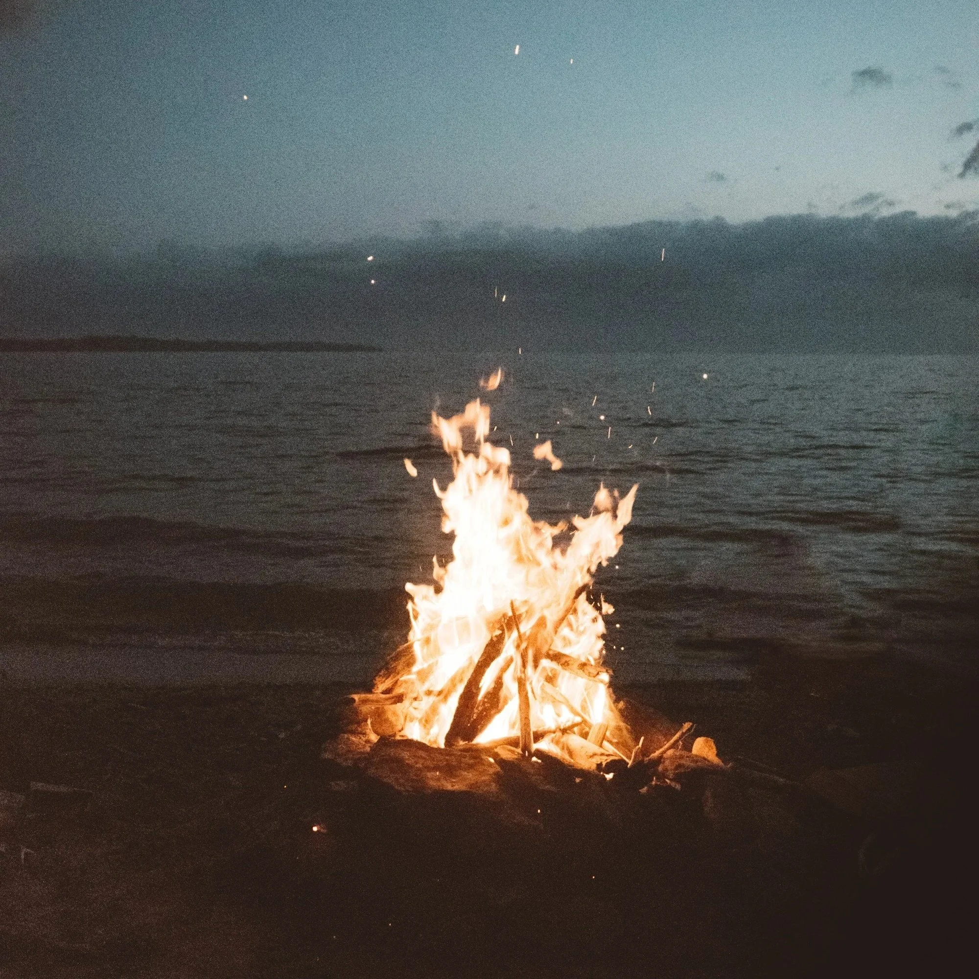 A bonfire on the beach with the ocean and cloudy sky in the background, taken during dusk or early evening.