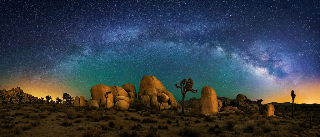 Nighttime desert landscape with large rock formations and Joshua trees under a star-filled sky with the Milky Way galaxy.