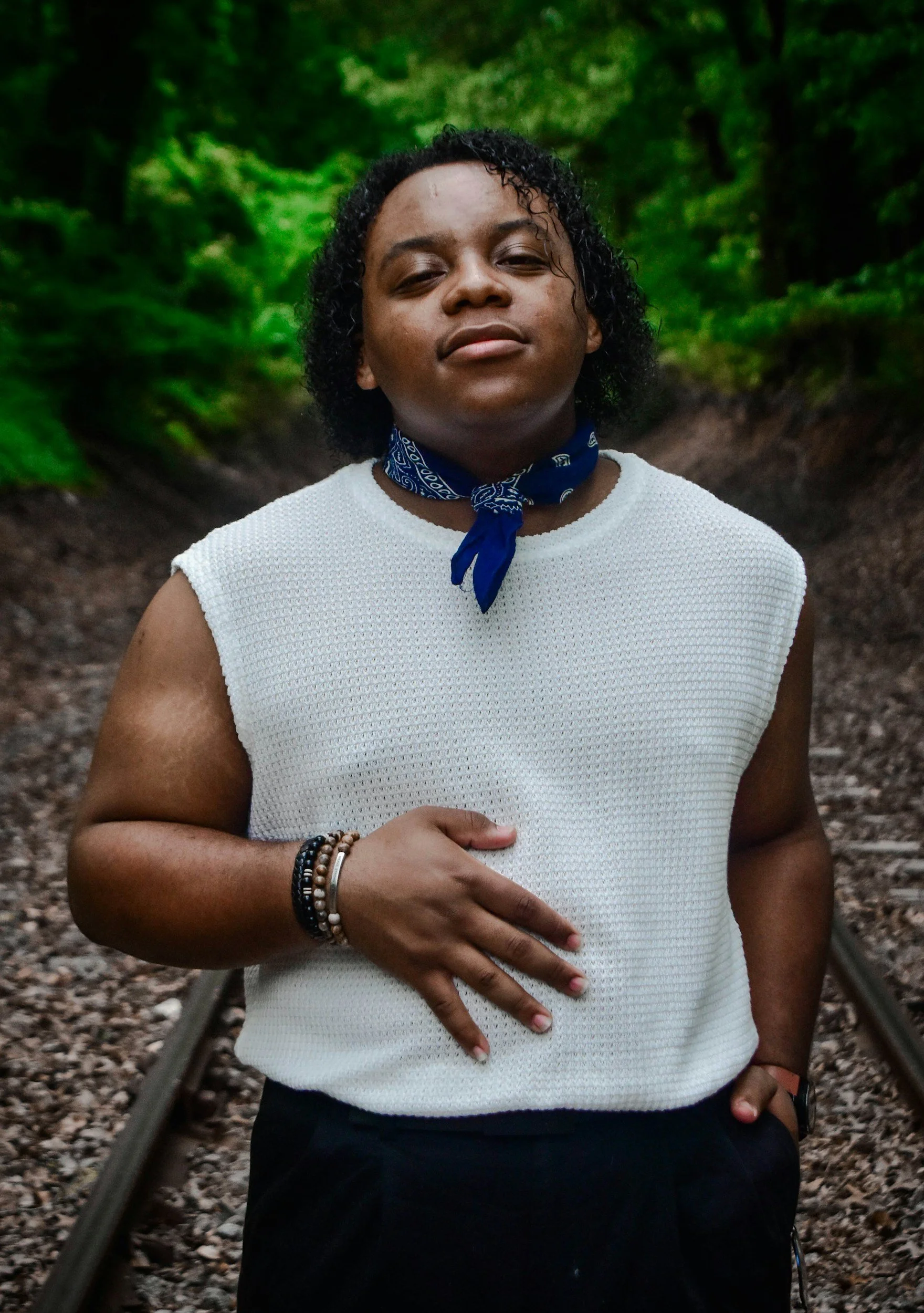 A person with short curly black hair wearing a white sleeveless top, a blue bandana around their neck, and bracelets on their right wrist, standing on railroad tracks in a green forest.