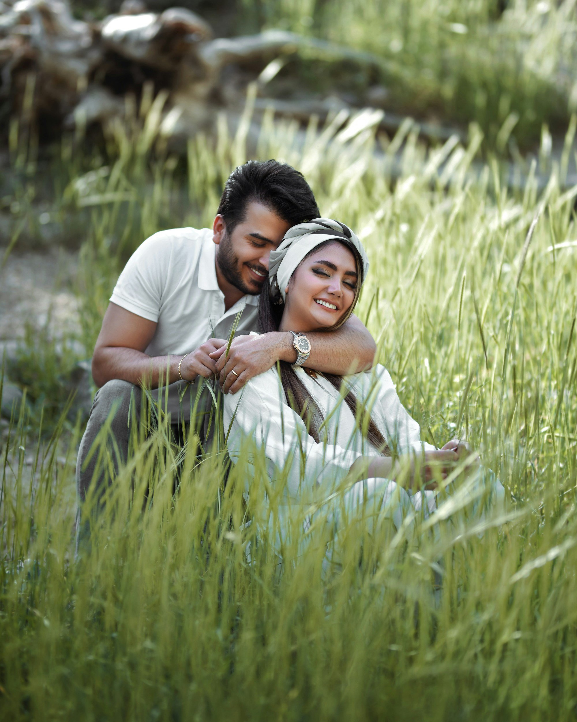 A smiling couple sitting in a lush green field near a body of water, with a background of trees and greenery.