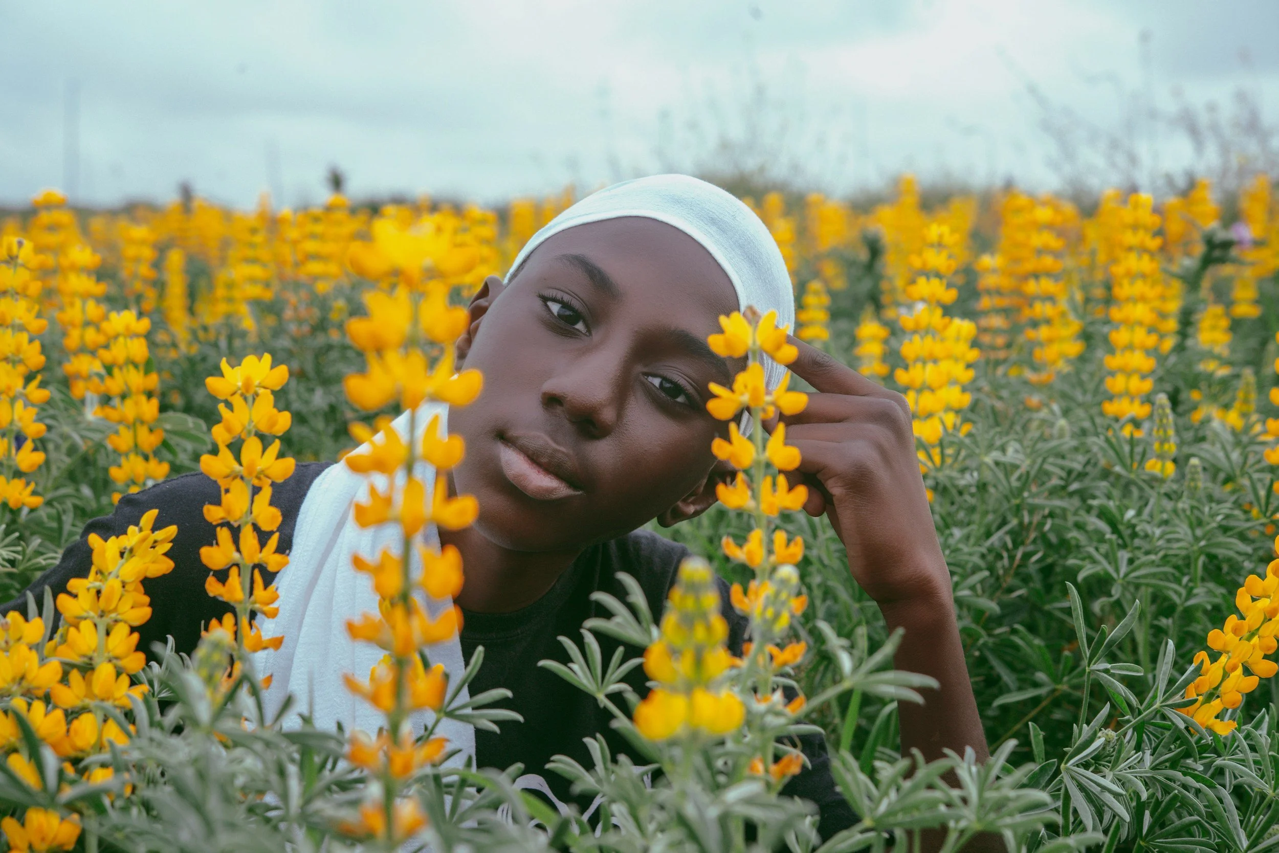 A person with dark skin, wearing a white headscarf and a black shirt with white accents, is among yellow flowers in a field. The person is leaning close to the flowers, touching their head with one hand and looking directly at the camera.