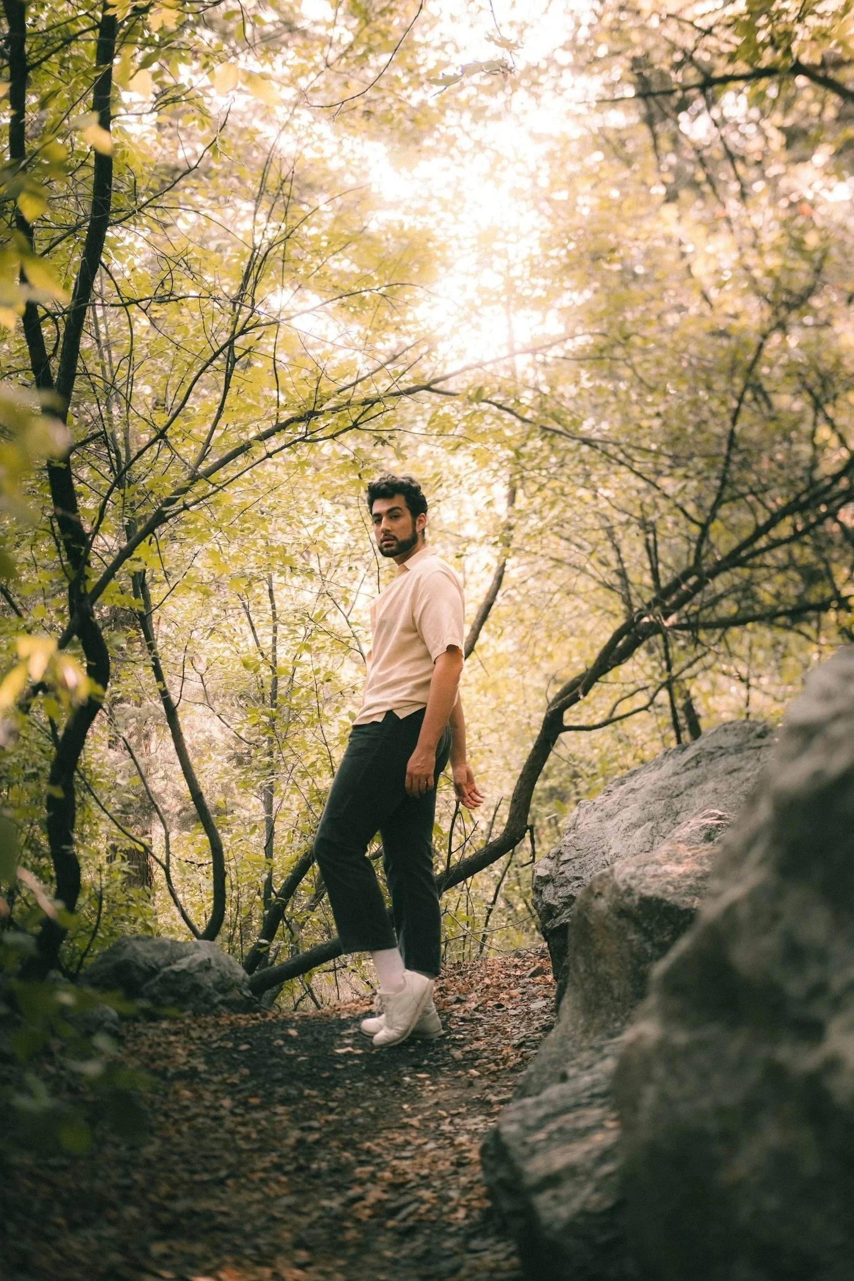 A young man with dark hair and a beard stands on a rocky trail surrounded by trees with green and yellow leaves, looking at the camera in a forested area during daytime with sunlight filtering through the foliage.