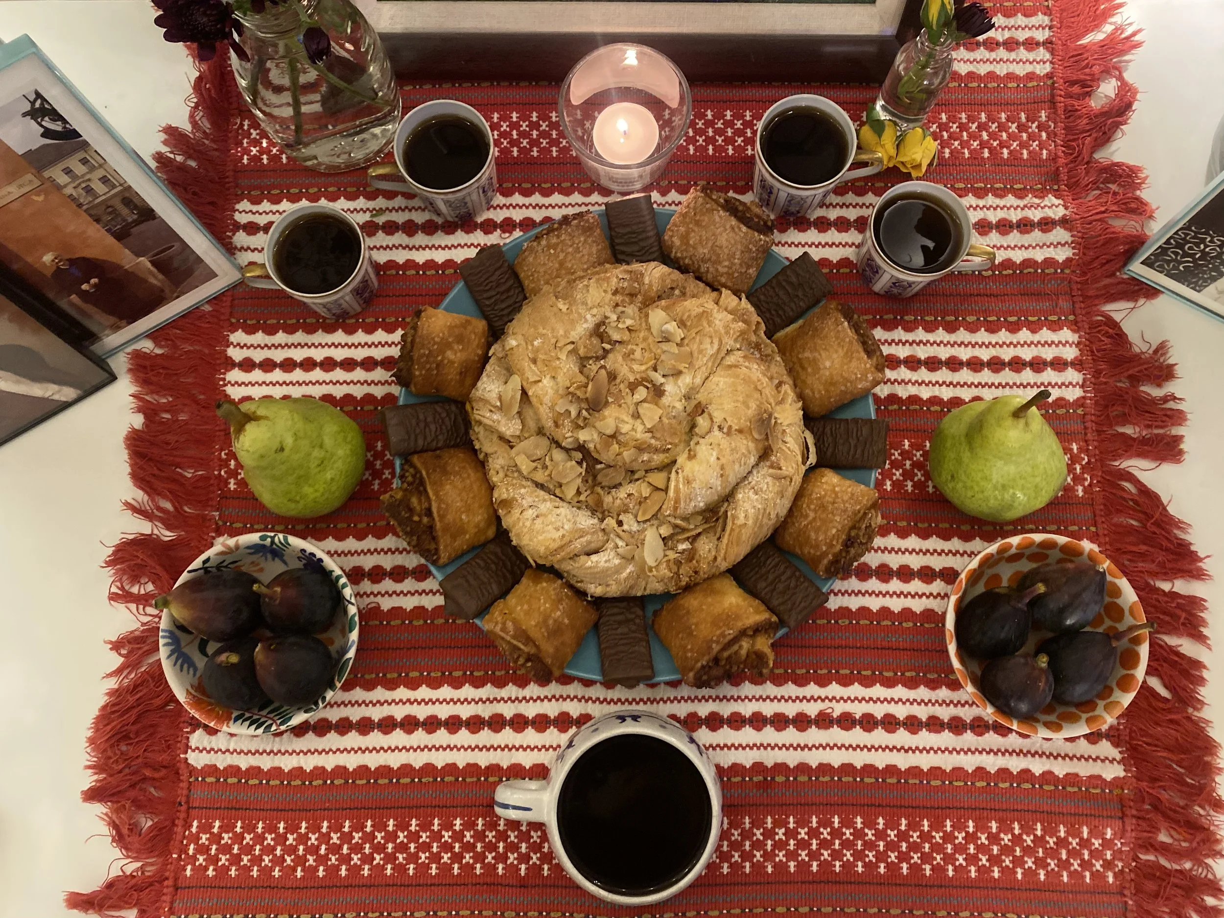 Table with a red and white woven traditional Hungarian tablecloth, featuring ancestral lineage healing offerings of pastries, coffee, fruit, flowers, a lit candle. This is part of divination and ritual healing.