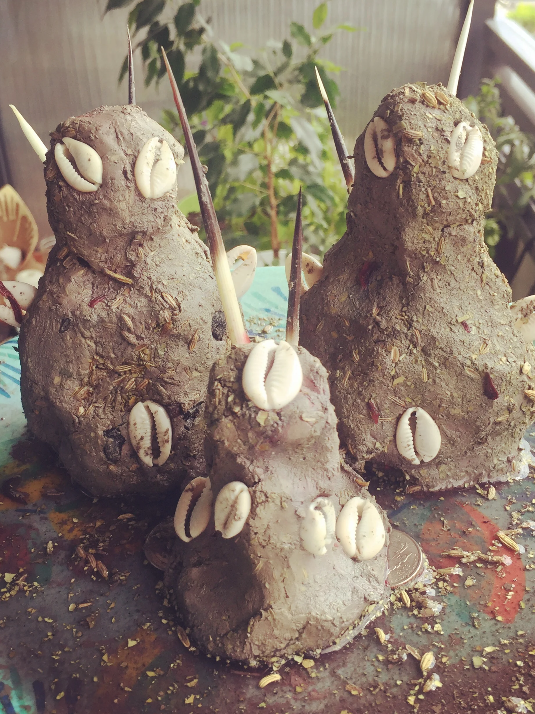 A shrine with three wedeme beings made of earth, decorated with white seashells, placed on a multicolored surface, with green plants and blurred background.