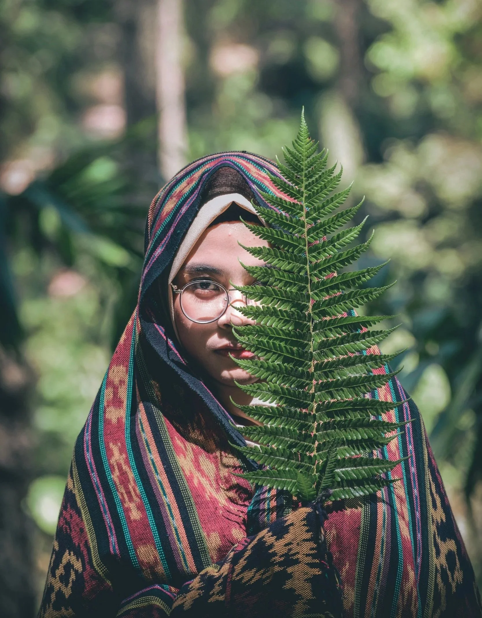 A person wearing glasses and a colorful, striped headscarf with a large green fern leaf partially covering their face, standing outdoors in a blurred forest background.