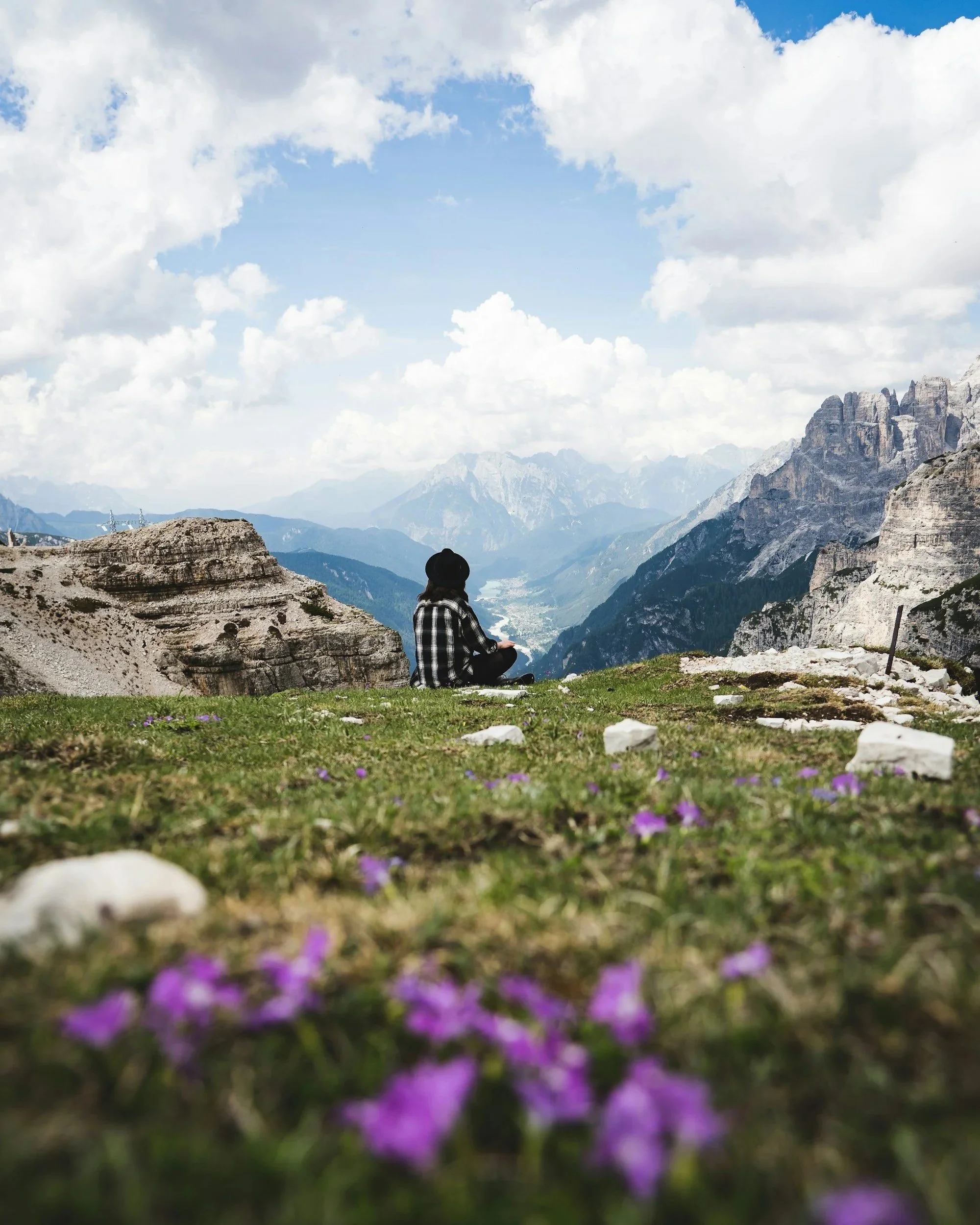 A person sitting cross-legged on a grassy area with purple flowers, overlooking a valley and mountain range under a partly cloudy sky.
