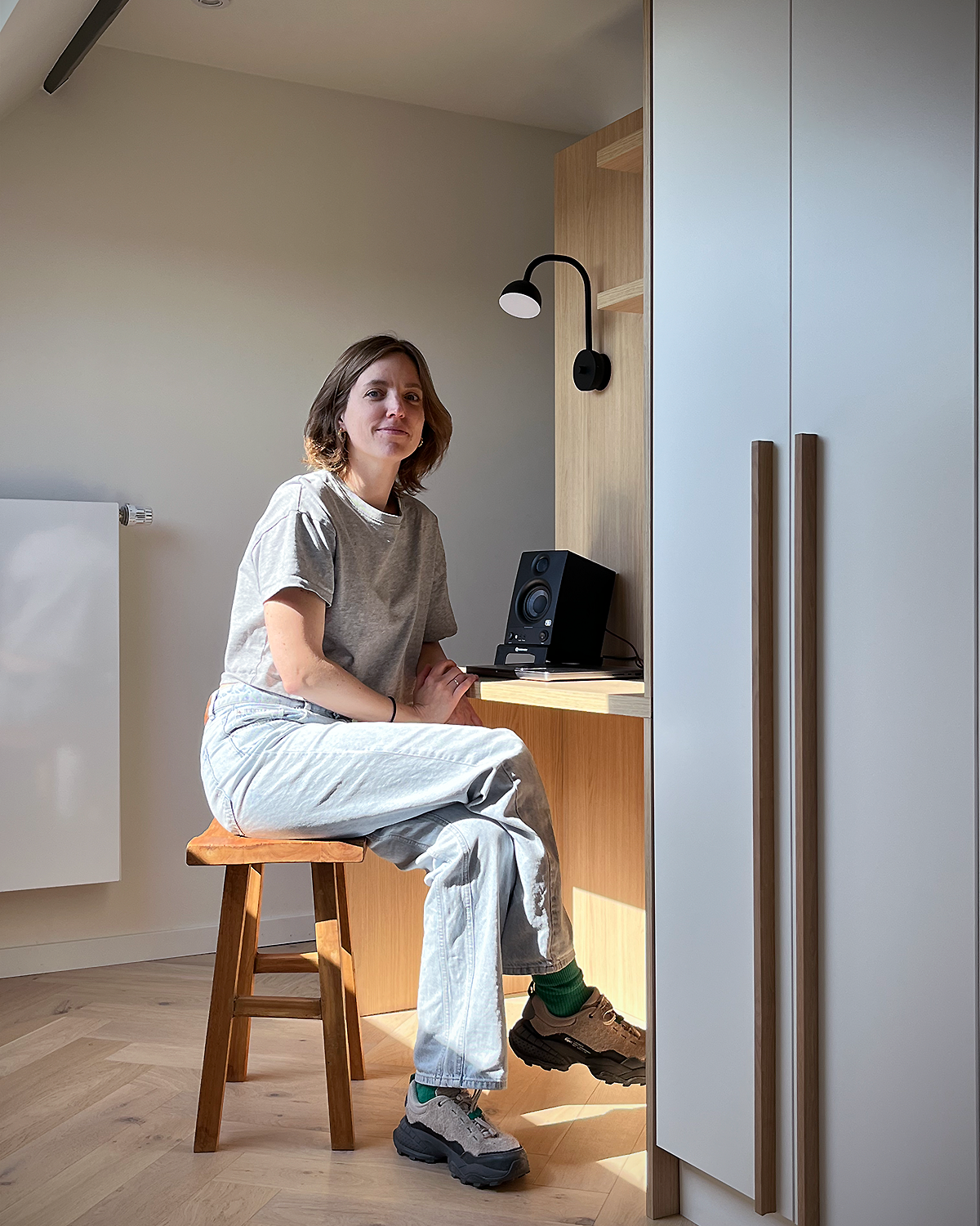 A woman sits on a wooden stool in a modern room, looking at the camera. She is wearing a gray t-shirt, light-colored jeans, and hiking shoes. A black speaker and a lamp are on a wooden shelf behind her, with a white cabinet to the right.