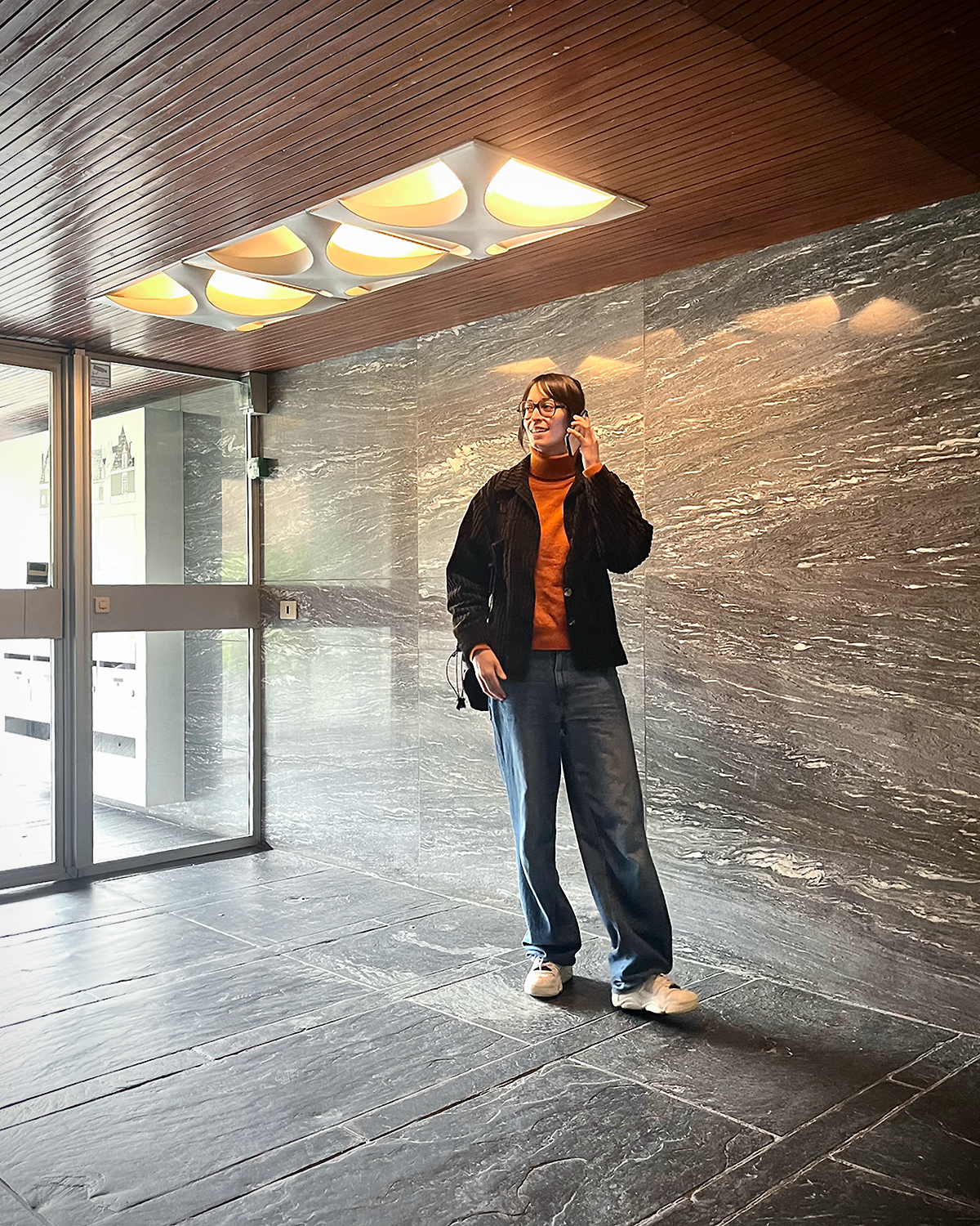 A woman standing indoors near a gray marble wall, talking on her cellphone, wearing glasses, a black jacket, orange shirt, blue jeans, and white sneakers.