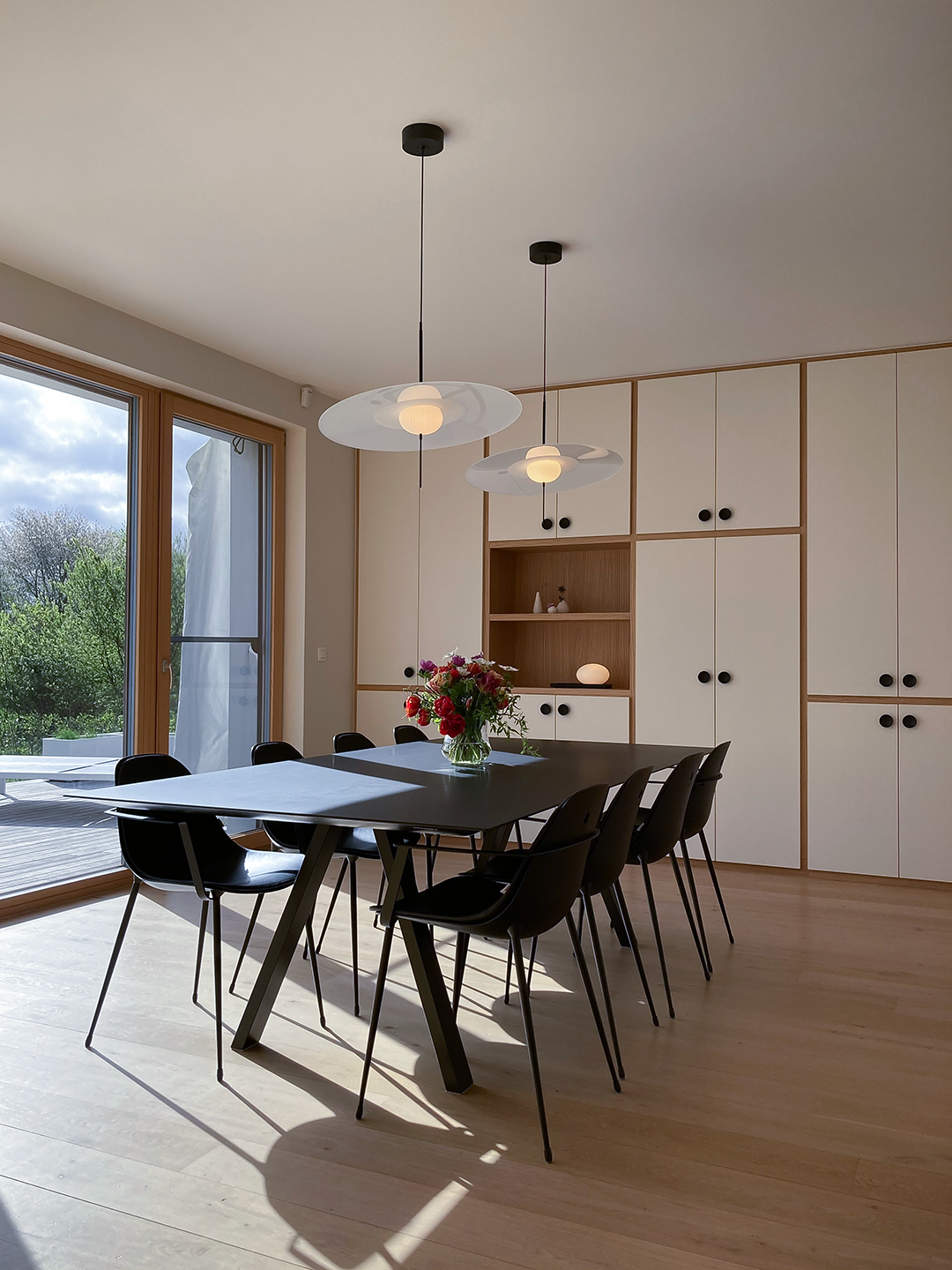 Modern dining room with black table, eight black chairs, a bouquet of flowers, large window with outdoor greenery, white cabinets, and two pendant lights with white shades.