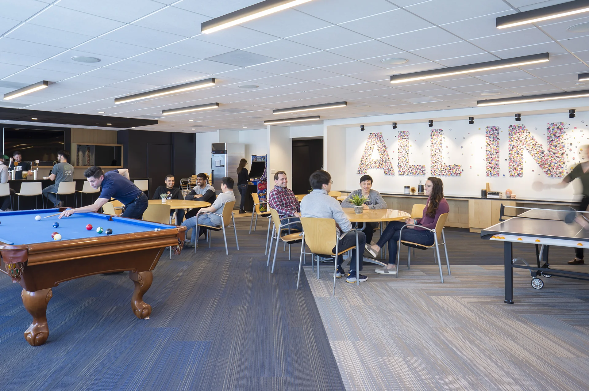 A photo of the game room - a pool table, ping pong table, arcade game, people sitting at tables, a bar, and a mural that reads “all in”