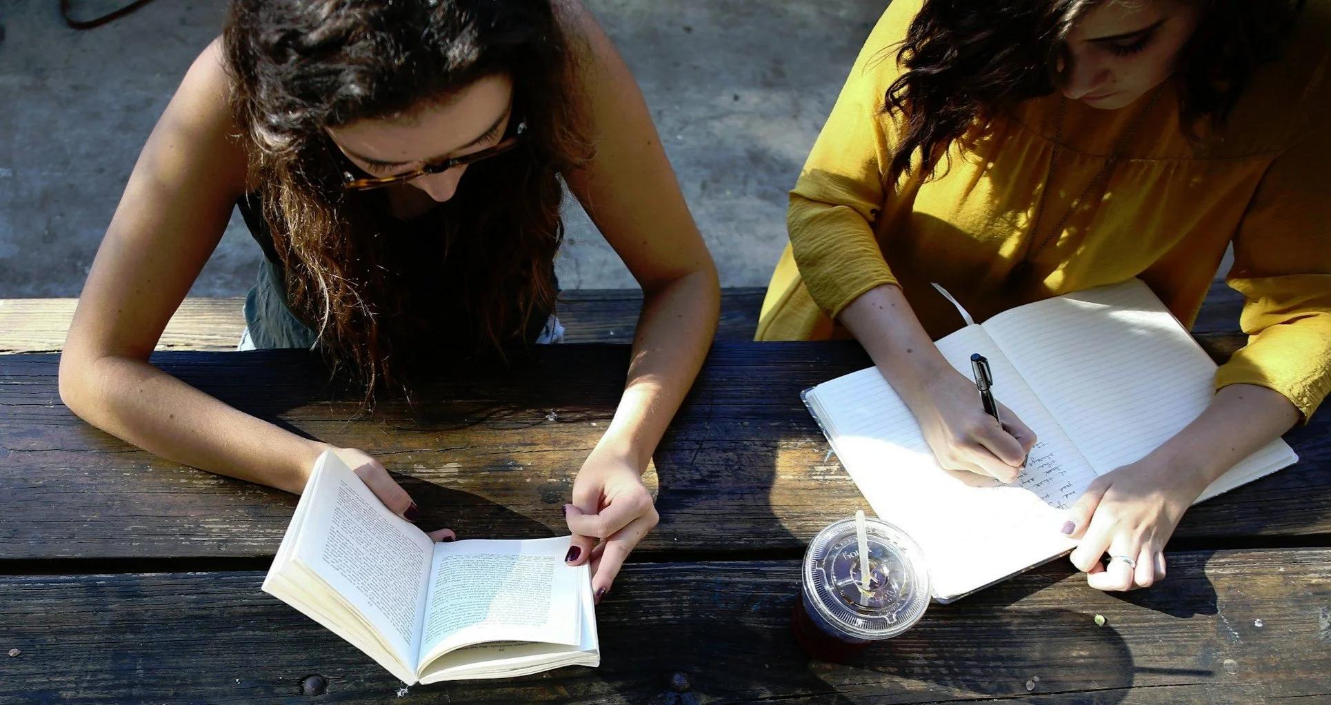 Two women sitting at a wooden picnic table outdoors, one reading a book and the other writing in a notebook, with a cup of iced coffee between them.