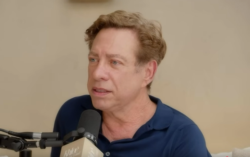 A man with light brown, wavy hair speaking into a microphone during an interview or podcast in a neutral room.
