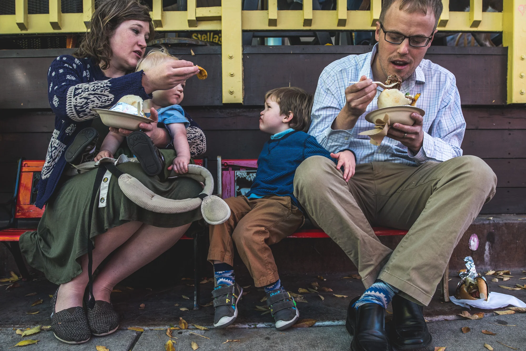 Family is sitting on a bench while mother tries to feed her son who doesn't want to eat what she's giving him, Pearl Street Farmer's Market, Denver, CO