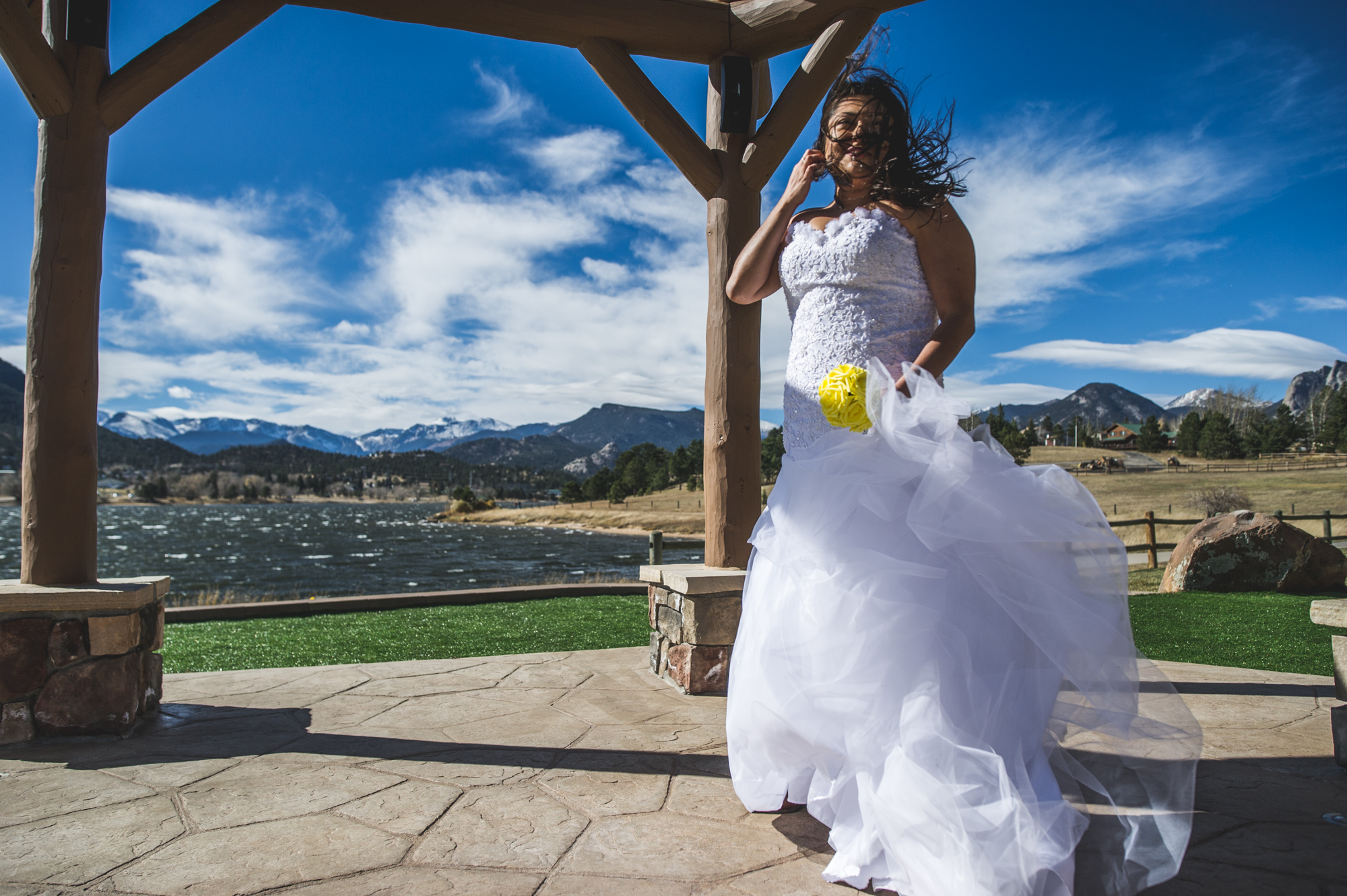 Bride stands in a gazebo at a resort in Estes Park, her hair and dress blowing wildly in the wind, color