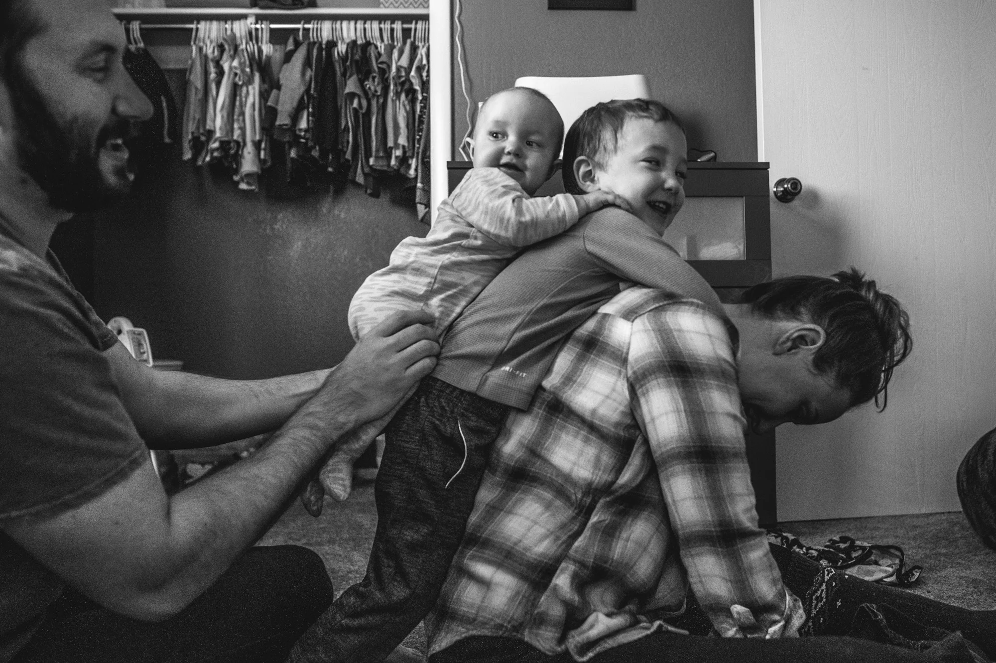 A woman bows her head as her 3 year old son hangs off of her back and her husband places their infant daughter on the back of their son, black and white, Denver, Colorado