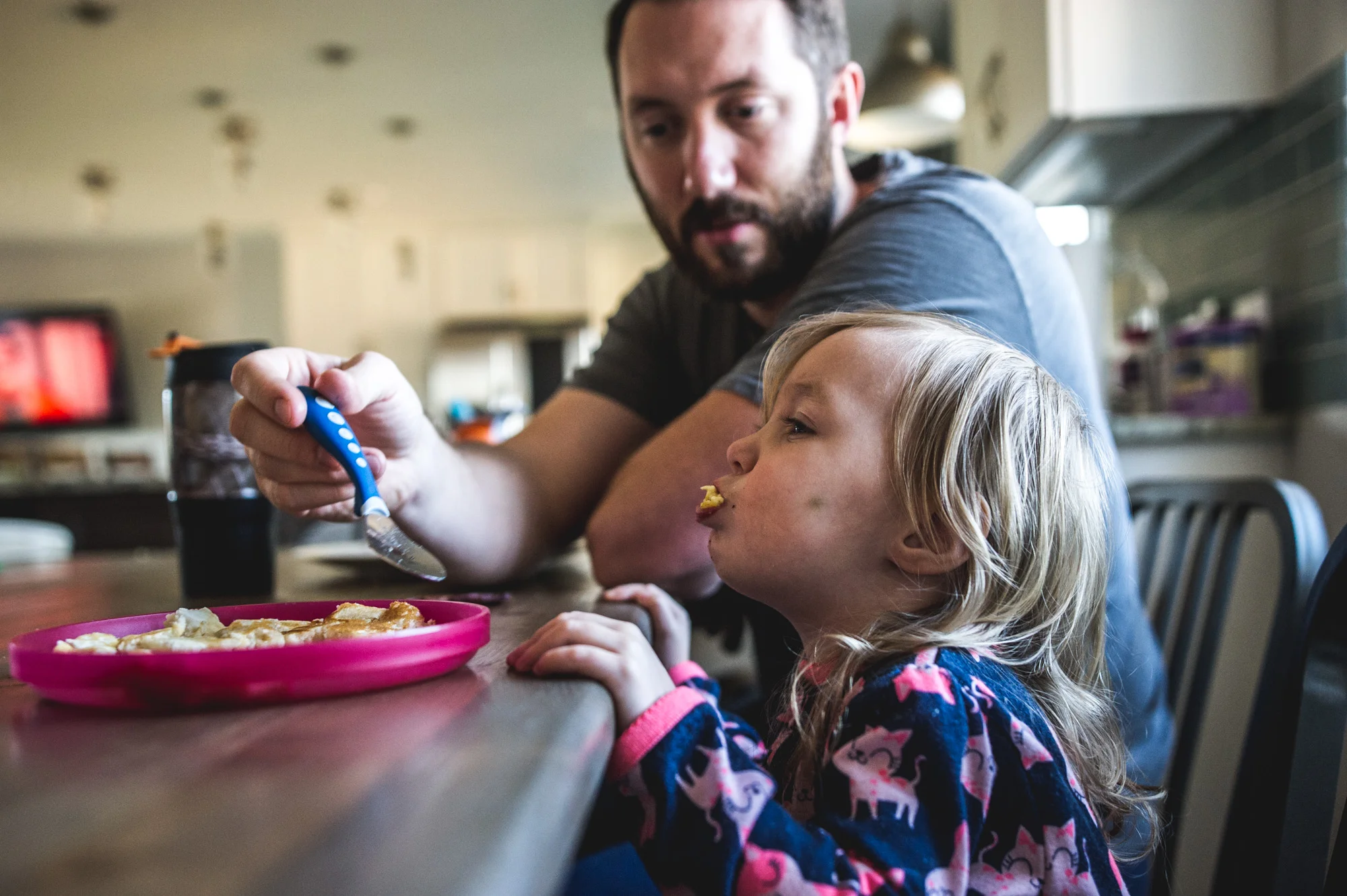 Little girl with eggs overflowing from her mouth as her father looks on, holding a spoon during their documentary photo session in Denver, Colorado