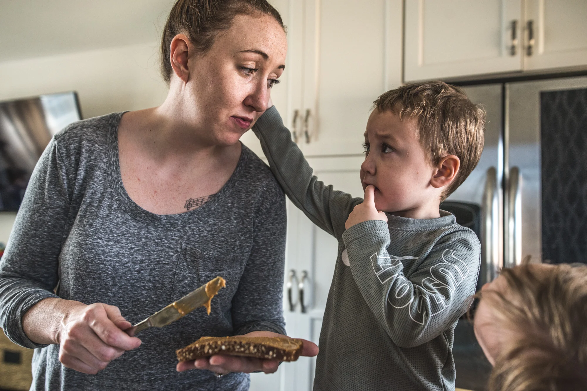 Mother spreads peanut butter on a piece of bread as her 3 year old son reaches up and touches his mother's face, color, day in the life session in Denver, CO