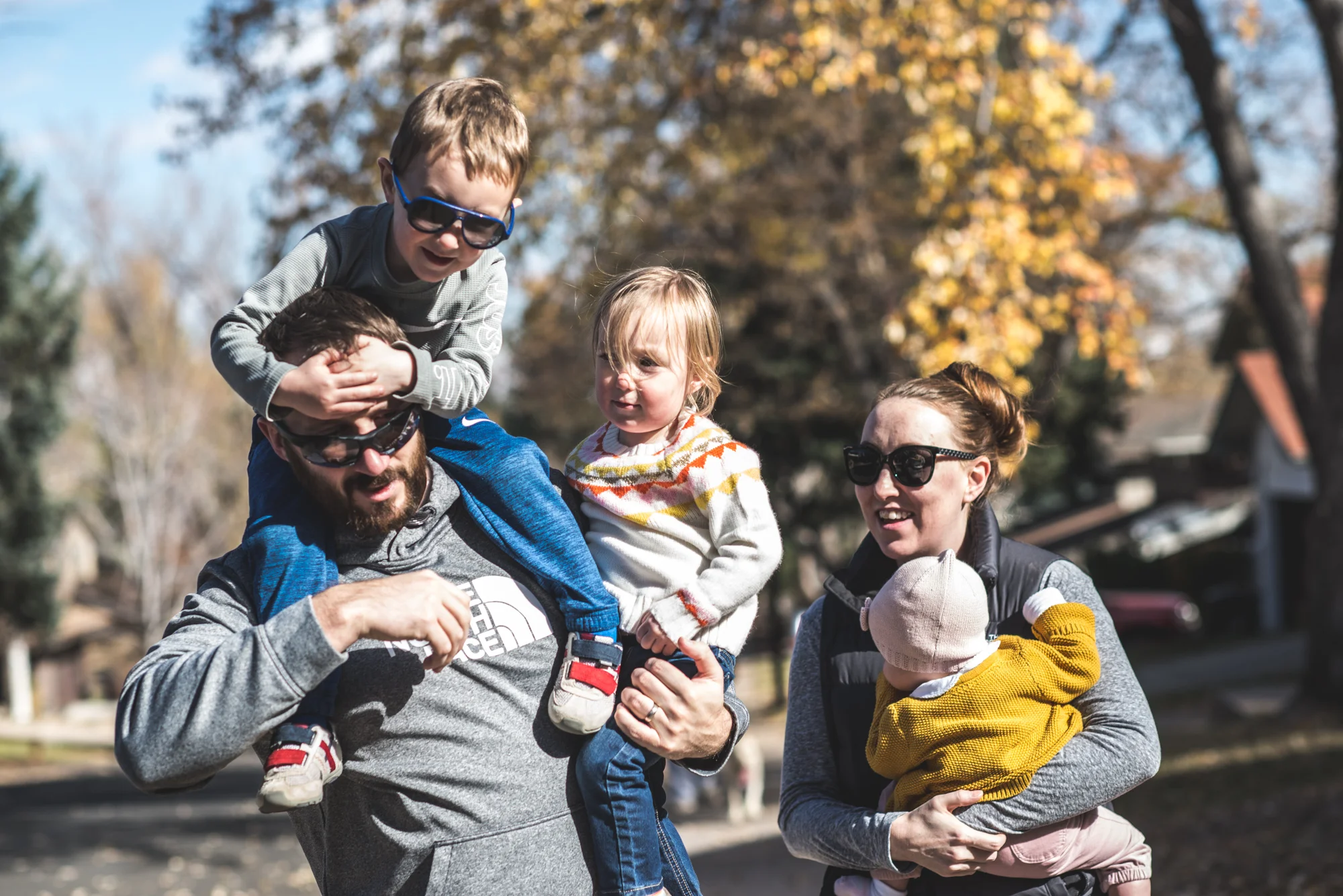 A father has his son on his shoulders and holds his daughter in his other hand while his wife walks beside him holding their 3rd child during a day in the life photography session in Denver, Colorado