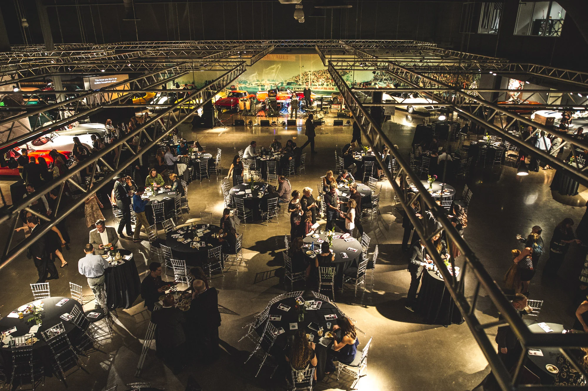 An overhead view of the Vehicle Vault main floor in Parker, Colorado. In the frame you can see the industrial supports overhead and tables filled with guests attending the Autism Society Winter Fundraiser. The walls are lined with vintage cars. Photo in color.