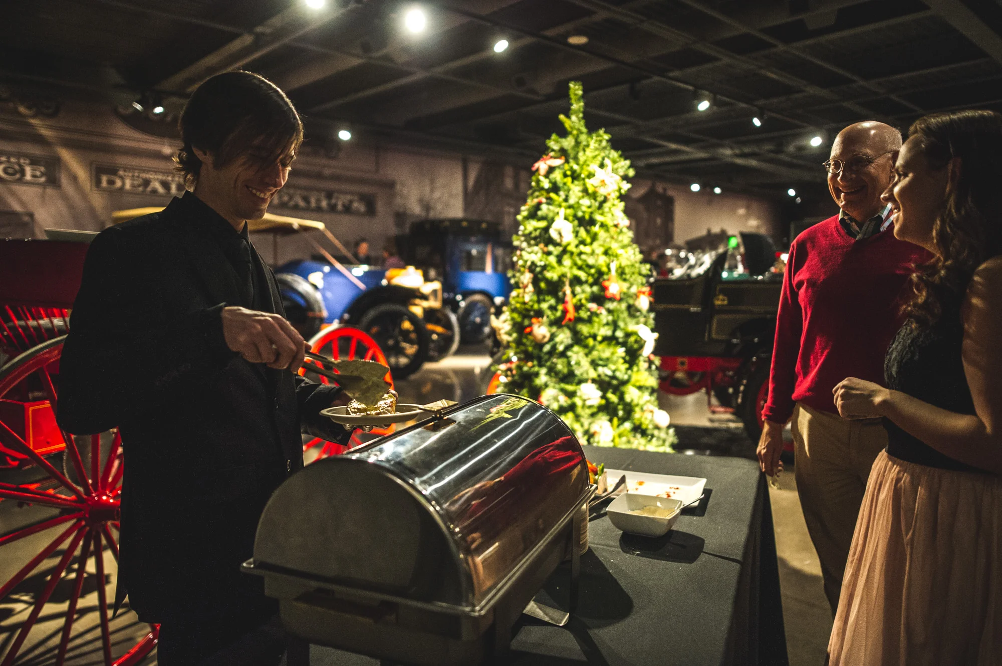 A man serves food to guests at the Autism Society Winter Fundraiser at the Vehicle Vault in Parker, Colorado. In the background are several vintage vehicles and a Christmas tree. Photo is in color.