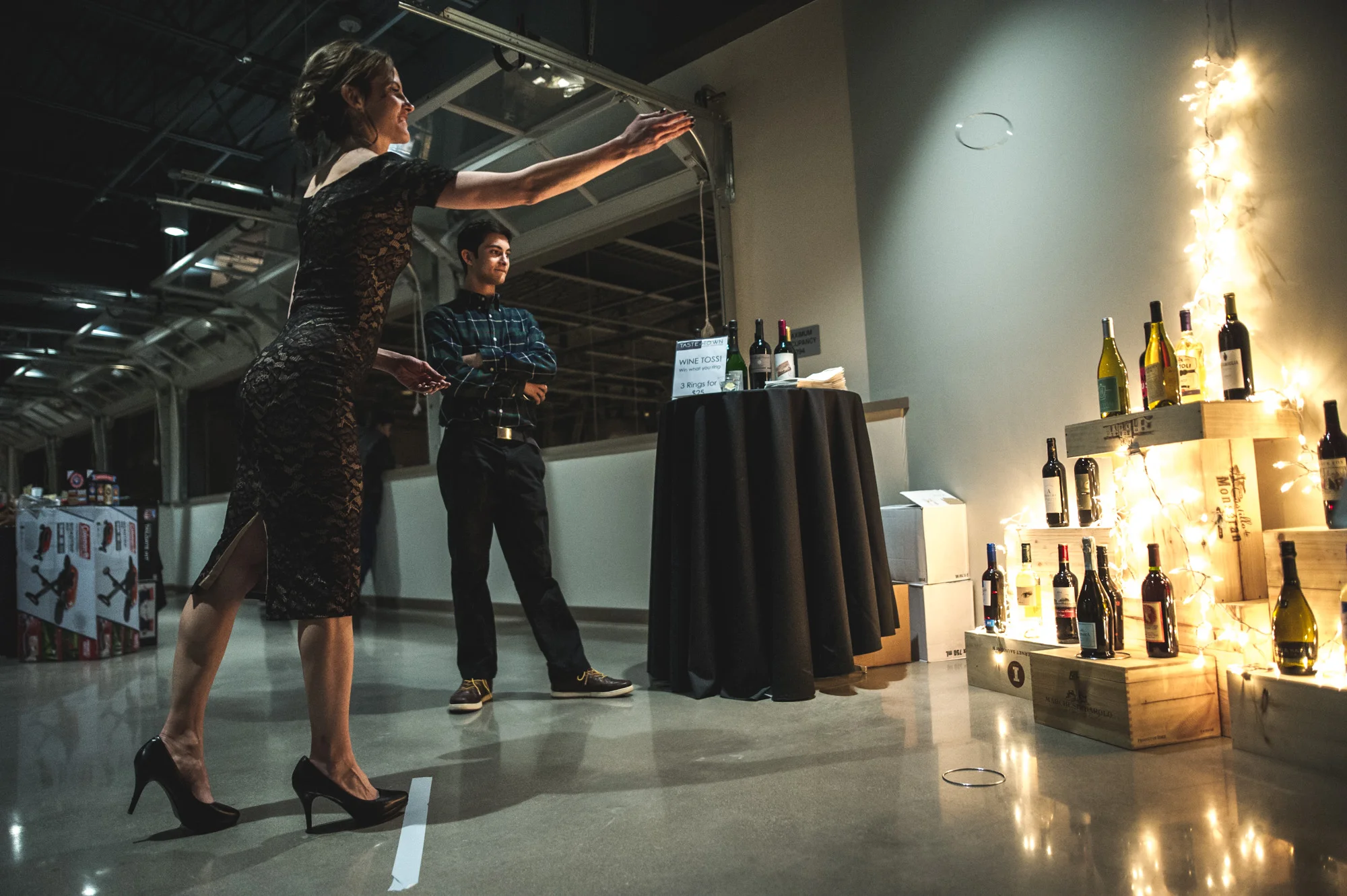 A woman in a cocktail dress and high heels throws rings at a wine display in hopes of getting one around a wine bottle as a man in the background watches. Photo in color.