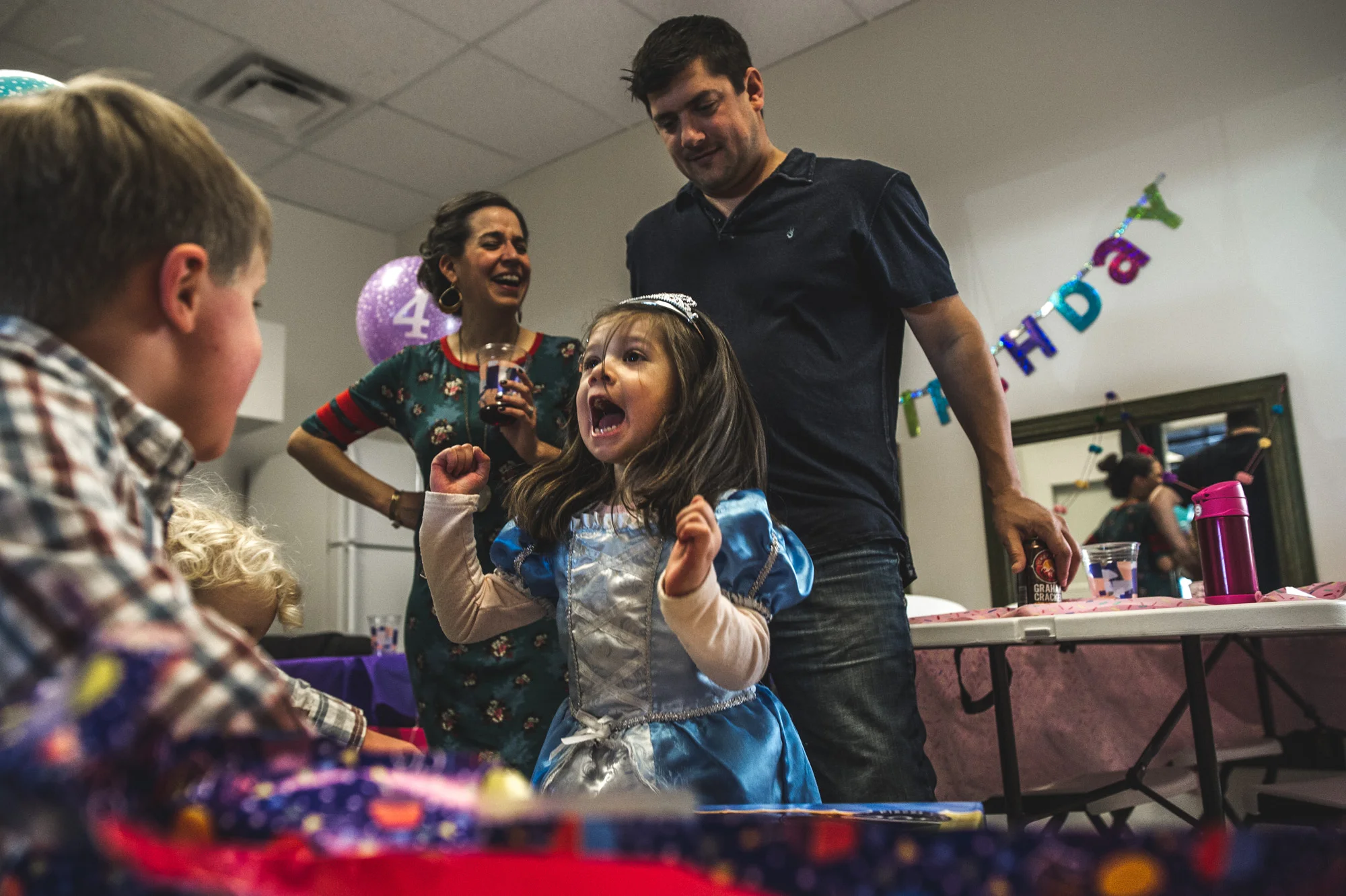 A little girl jumps for joy as her parents look on. The little girl is wearing a princess dress and crown and is celebrating her birthday at Family Connections kids' gym in Littleton, Colorado. Photo is in color.