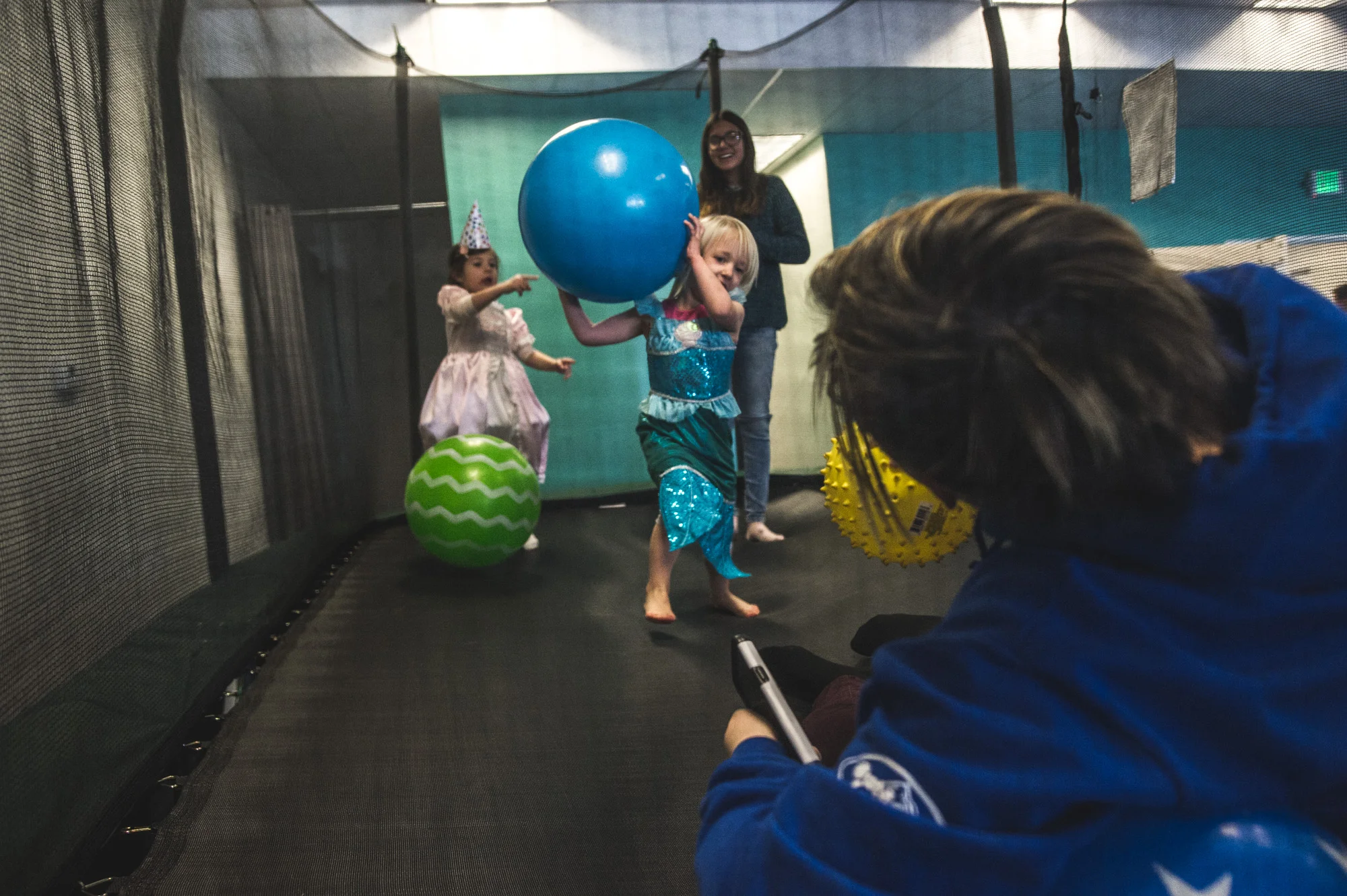 Four people are on a trampoline, 2 younger kids and 2 bigger kids, and one of the smaller children takes aim at one of the bigger kids with a large blue ball at a kids' gym in Littleton, Colorado.