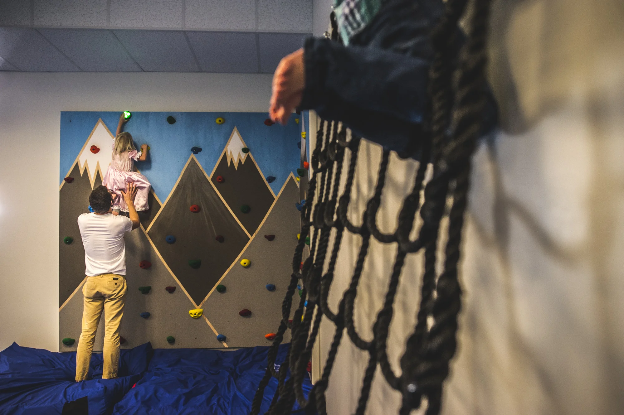 A young girl climbs a rock wall with the help of her dad while wearing a pink princess dress. In the foreground to the right can be seen the legs of a little boy climbing a net. Littleton, Colorado.