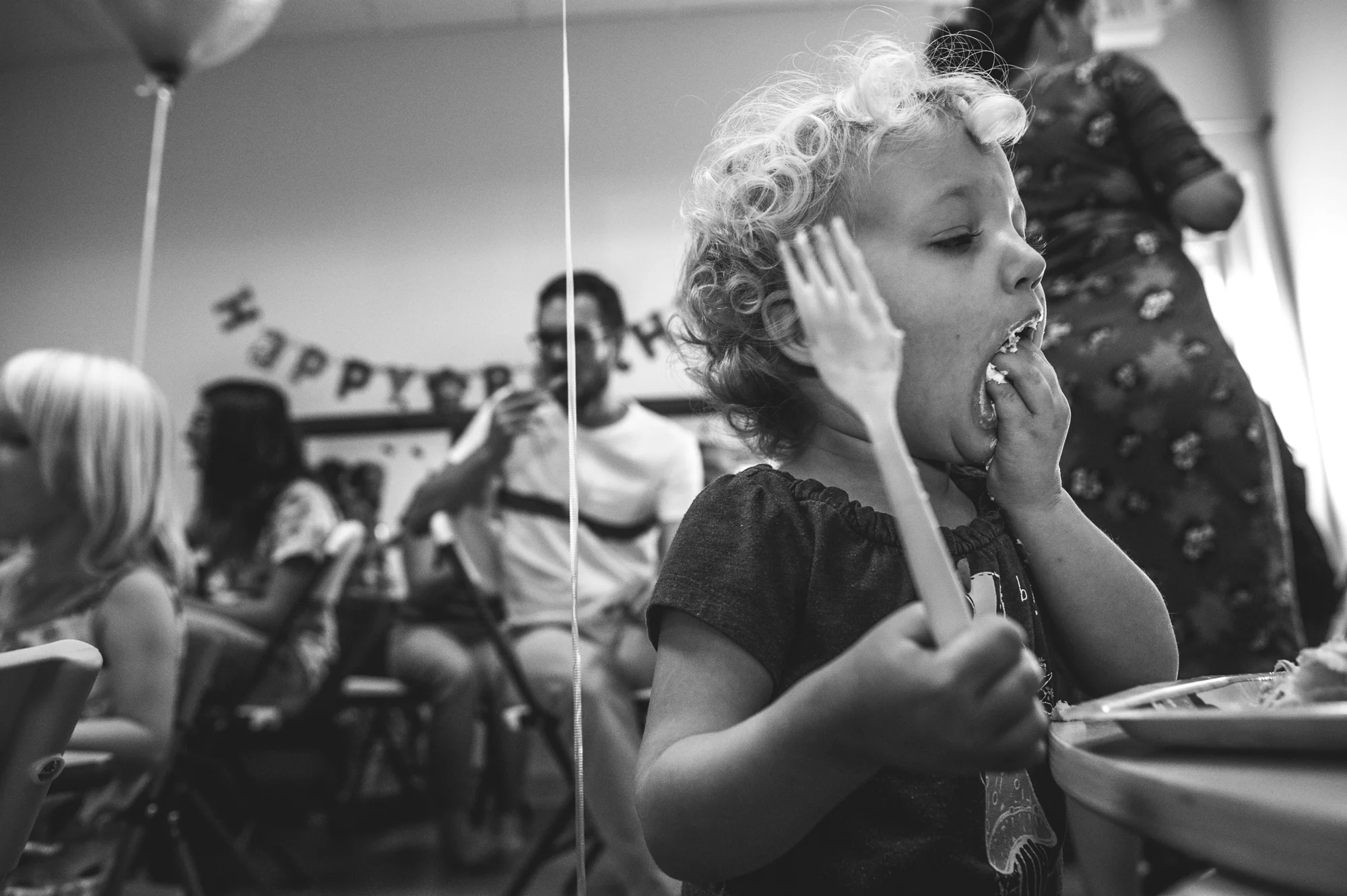 A little girl shoves a bunch of cake in her mouth using her hand despite having a fork in the other hand, photo in black and white